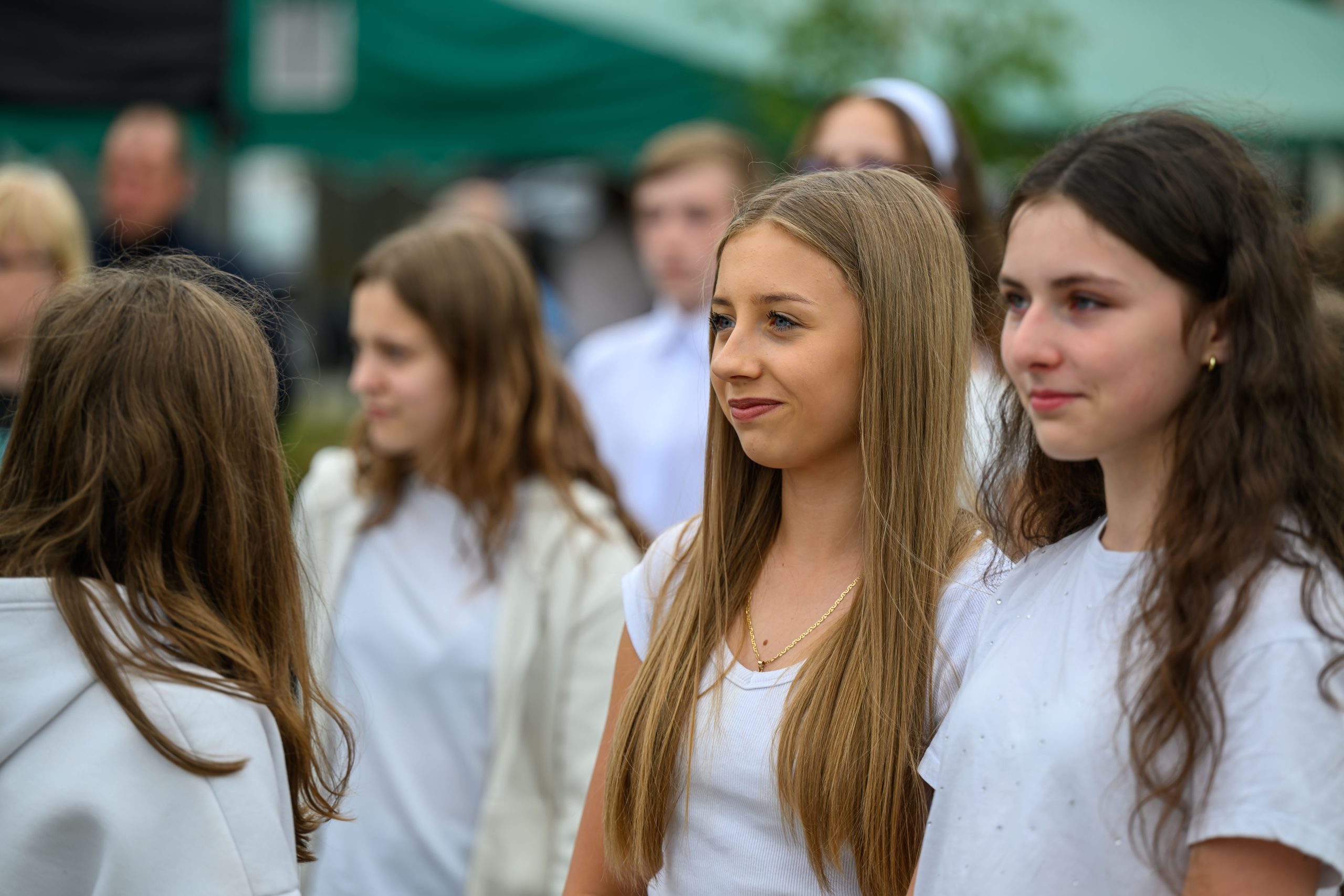 Na zdjęciu widać kilka młodych osób na zewnątrz, ubranych na biało. Tło jest rozmyte z zielonymi i ciemnymi elementami. Dominują naturalne kolory i subtelne uśmiechy.