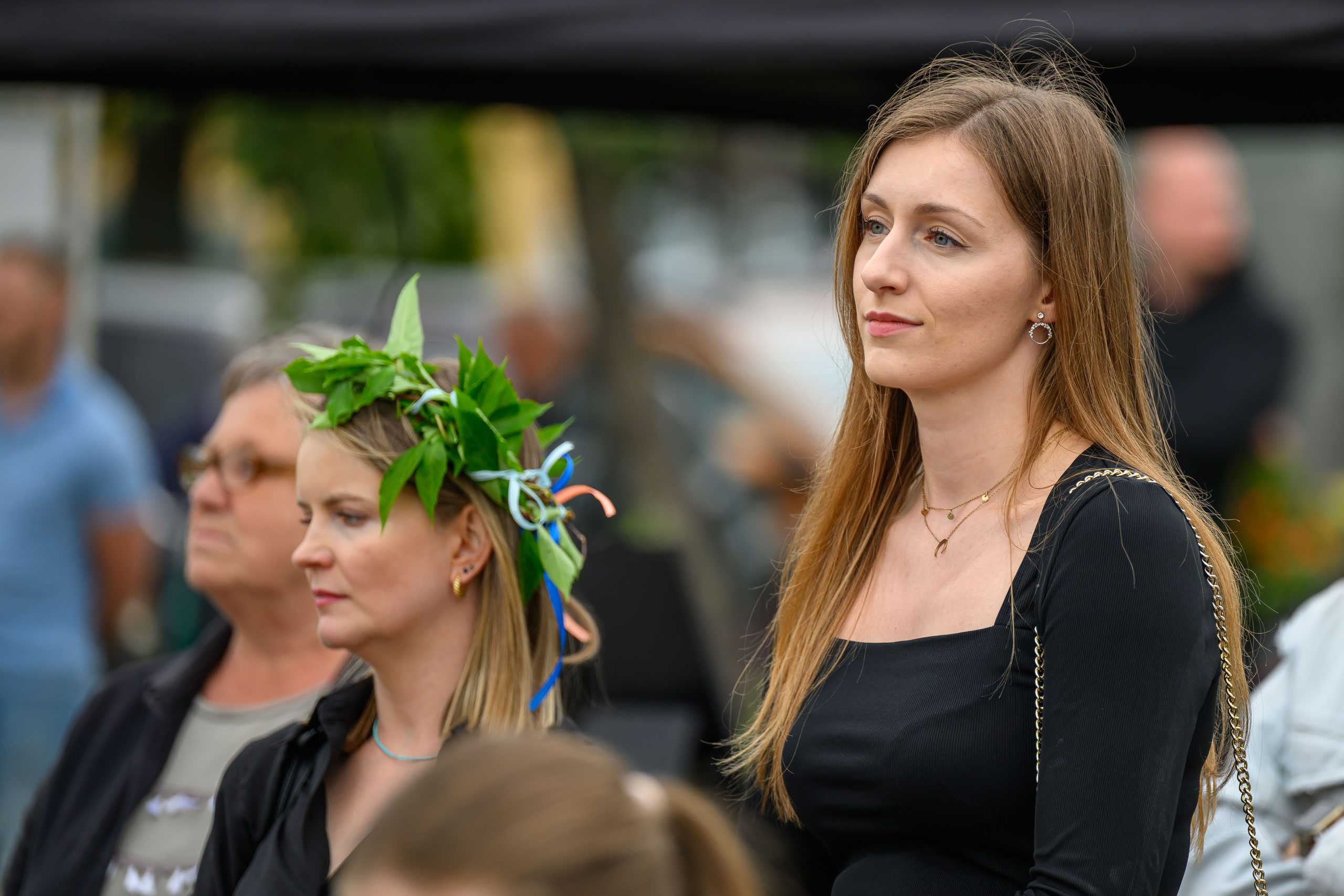 Na zdjęciu są dwie kobiety, jedna w wianku z liści, druga w czarnej sukience. W tle rozmyta grupa ludzi. Dzień jest pochmurny.