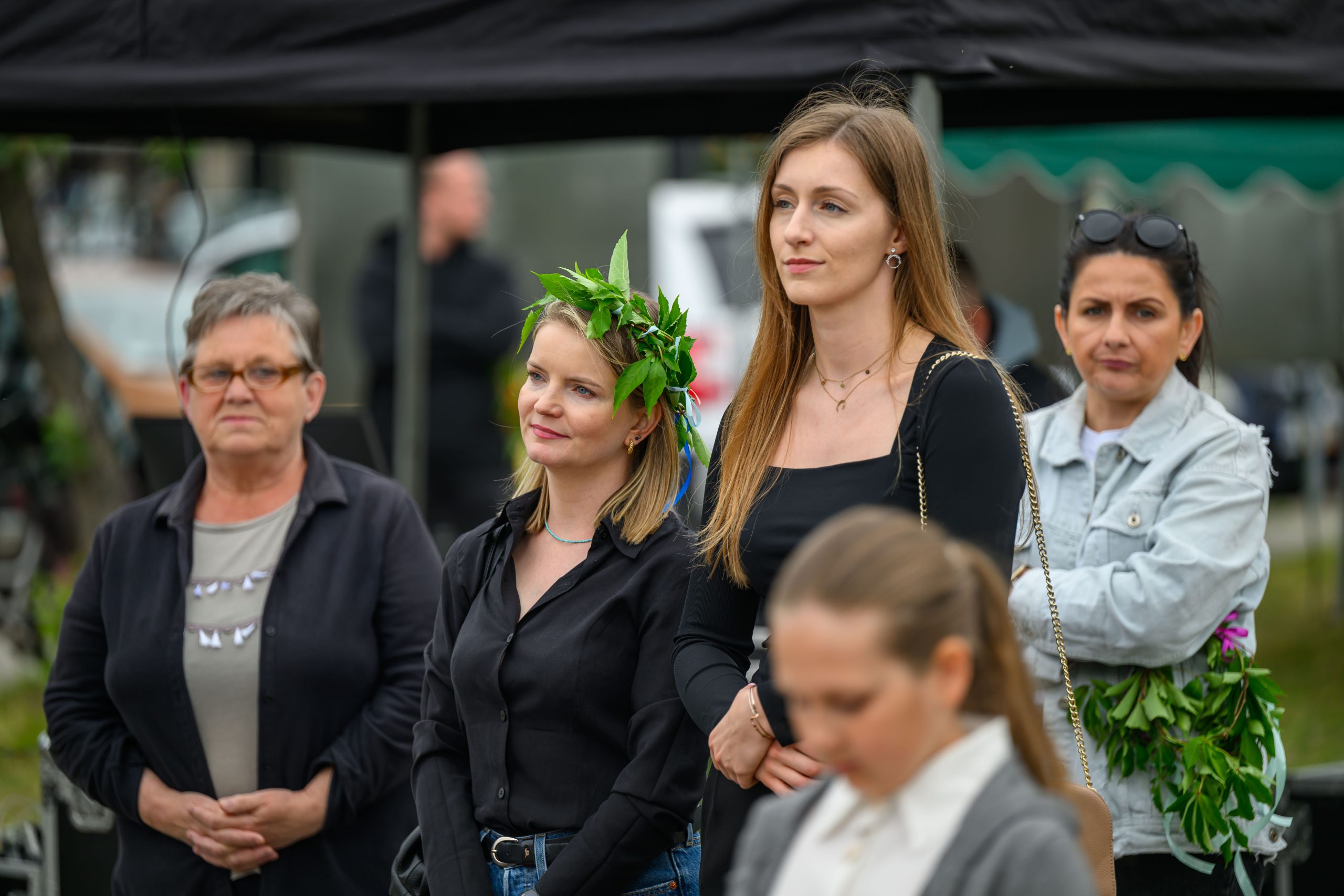 Grupa ludzi stoi na zewnątrz. Kobieta ma wianek z liści. W tle są namioty w ciemnych kolorach.