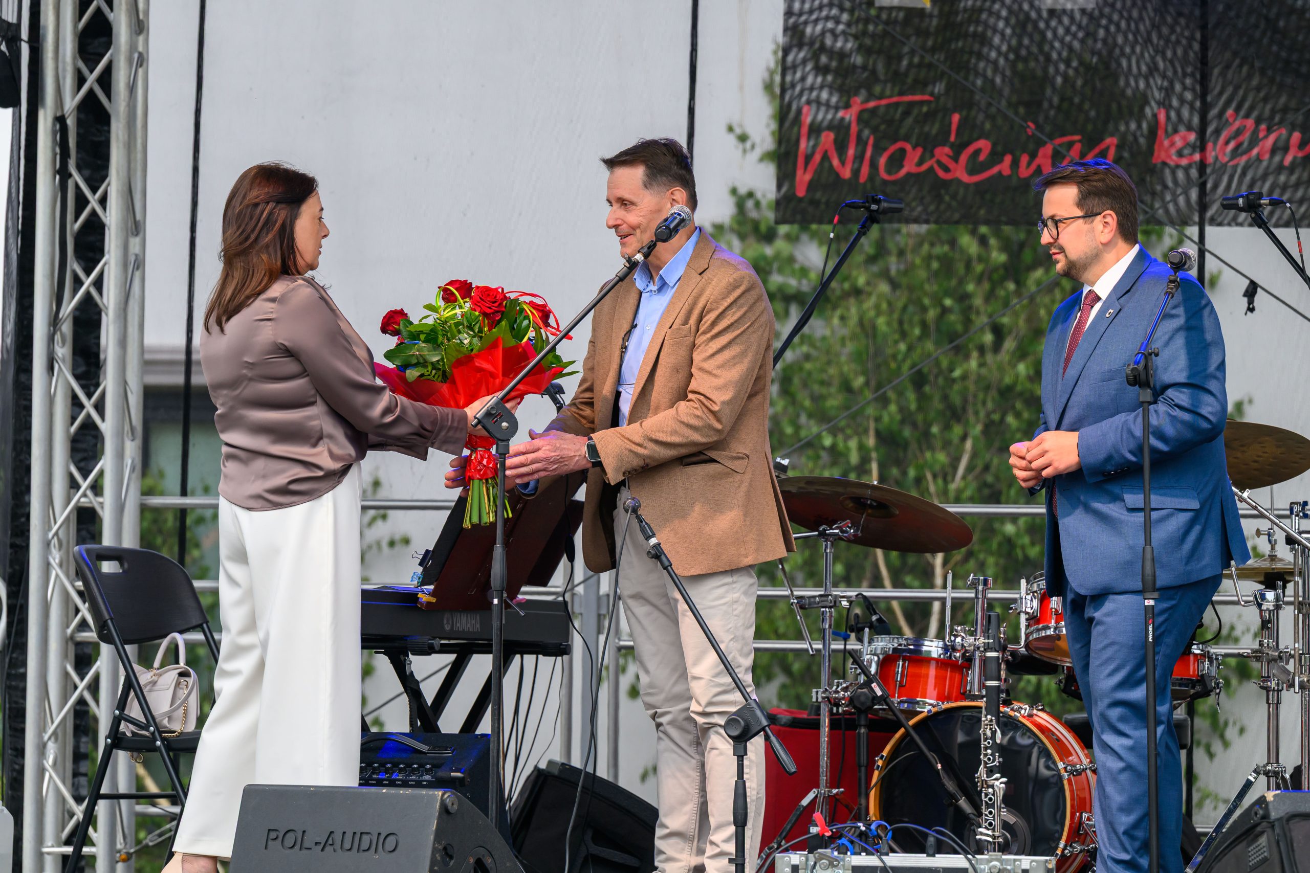 Trzy osoby na scenie, kobieta wręcza bukiet czerwonych róż mężczyźnie. Tło to sprzęt muzyczny i zielone drzewa. Strój elegancki.