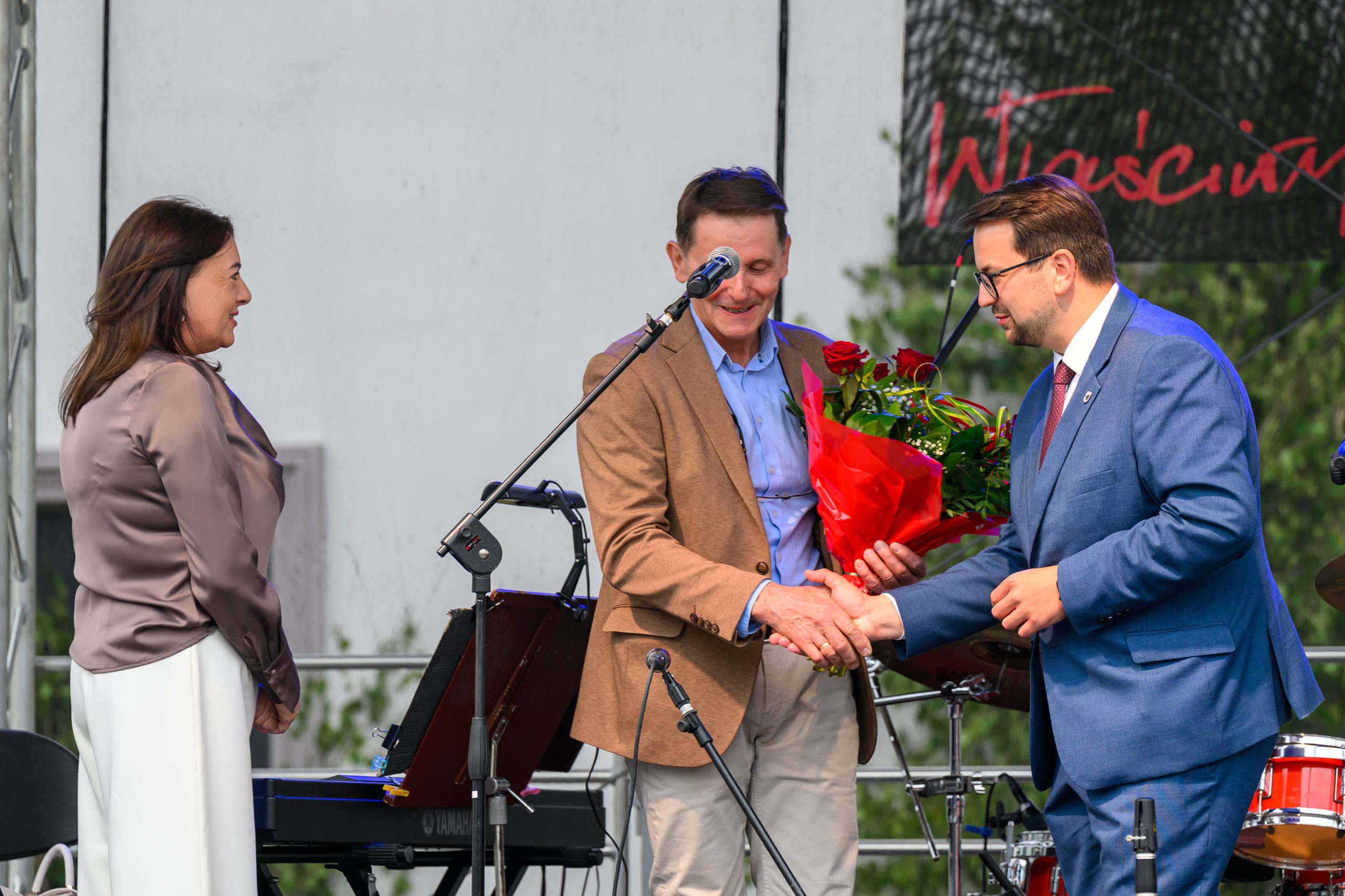 Trzech ludzi na scenie; mężczyzna w garniturze podaje dłoń drugiemu, trzymającemu czerwone róże. Kobieta patrzy z boku.