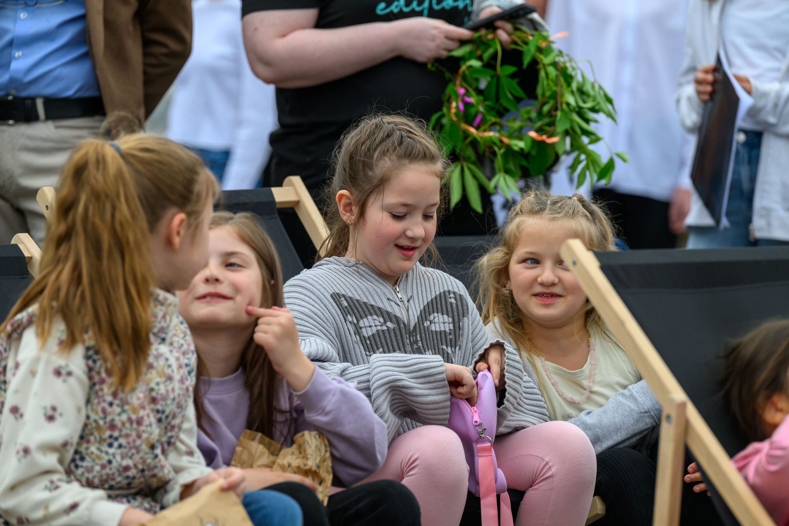 Grupa uśmiechniętych dzieci siedzi na leżakach na zewnątrz. Dziewczynki rozmawiają w swobodnej atmosferze. W tle widać osoby dorosłe i zielony wianek.