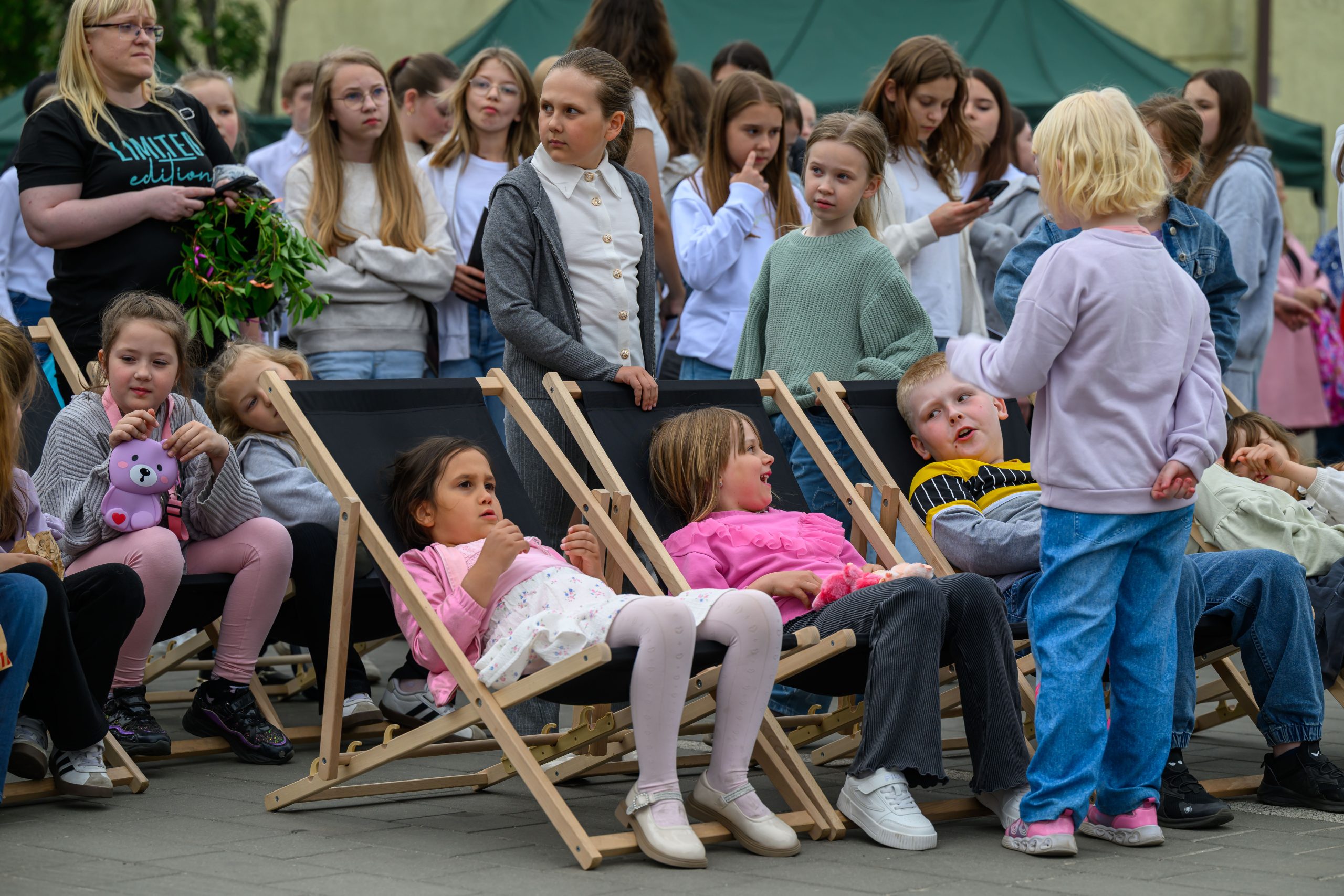 Dzieci siedzą na leżakach w kolorowych ubraniach, otoczone grupą rówieśników. W tle zielono-beżowy namiot. Scena na świeżym powietrzu.