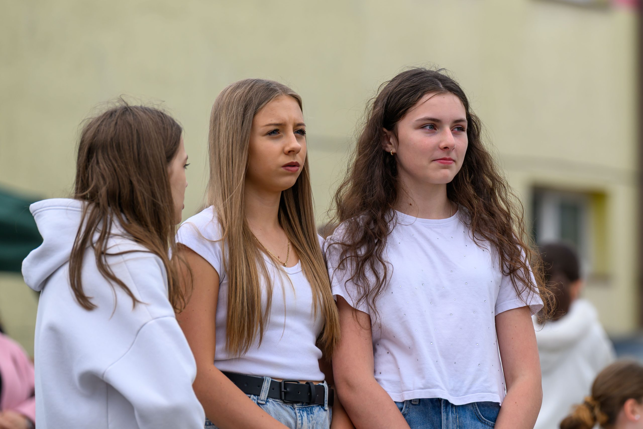 Trzy dziewczyny w białych koszulkach stoją na zewnątrz, na tle jasnej ściany. Przeważa kolor biały i beżowy. W tle widoczne są rozmyte postacie.