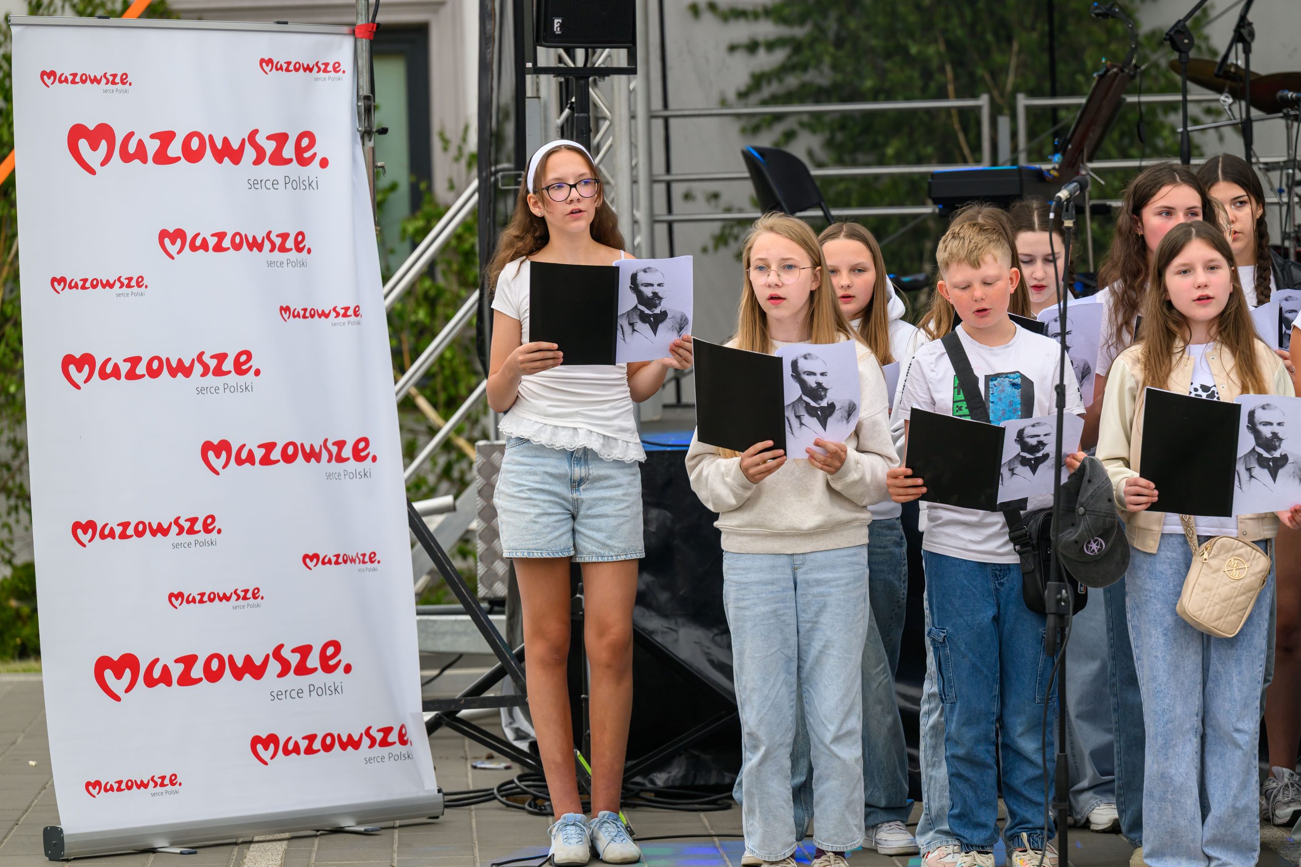 Grupa dzieci trzyma zdjęcia Piłsudskiego, stojąc przy banerze Mazowsze. Ubrane w jasne ubrania, śpiewają na zewnątrz. W tle rośliny i scena.
