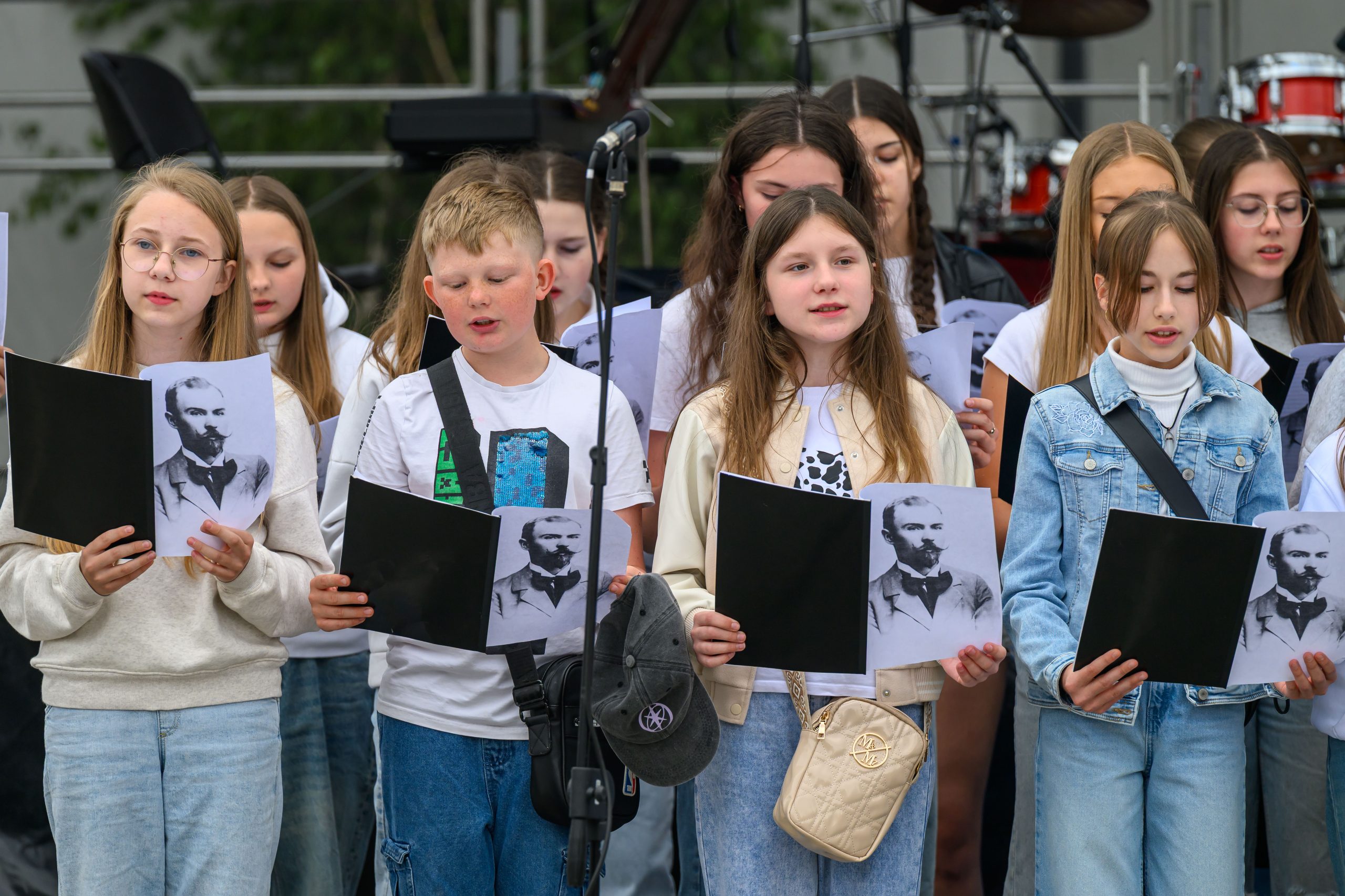 Grupa dzieci w białych bluzkach i dżinsach śpiewa na scenie, trzymając czarne teczki. W tle widoczne instrumenty perkusyjne. Dzieci mają przed sobą zdjęcia mężczyzny z brodą.