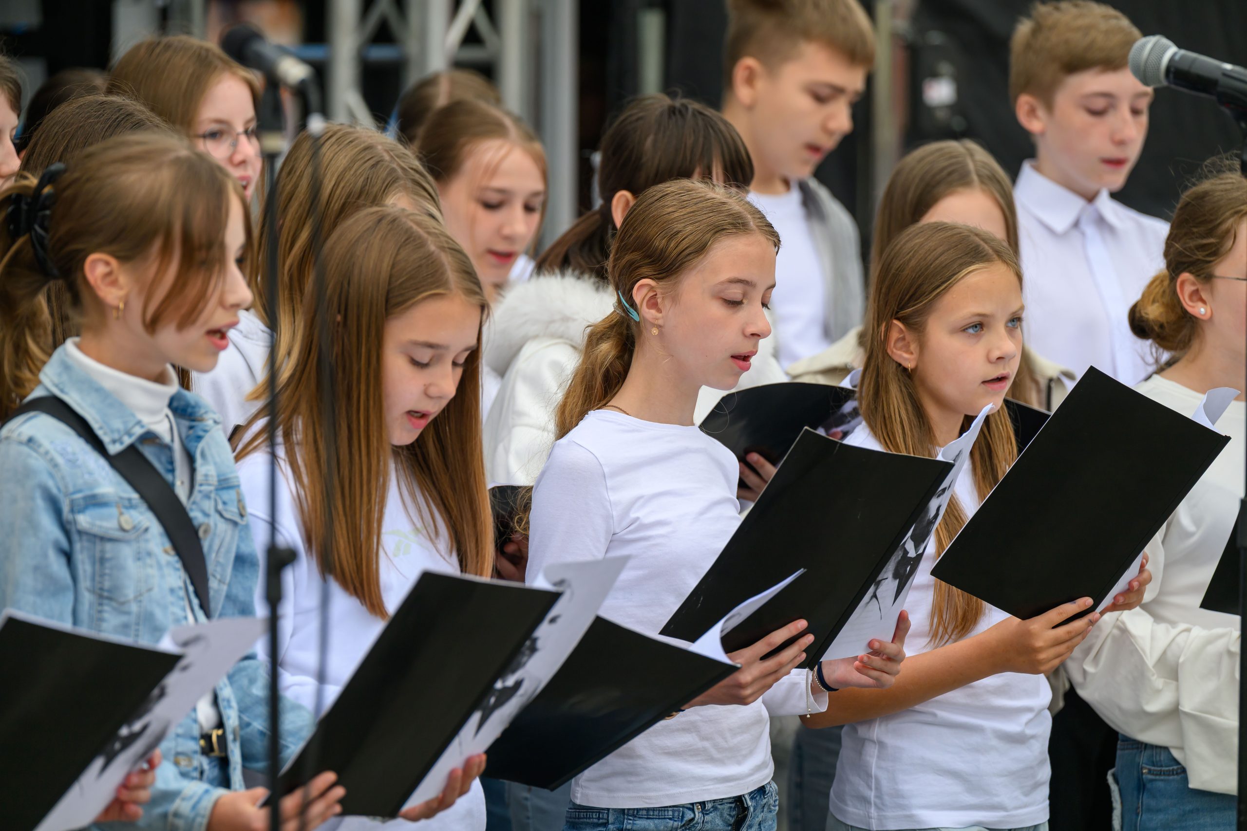 Grupa dzieci śpiewa, trzymając czarne teczki z nutami. Ubrane są w białe bluzki i stoją na tle mikrofonów. Scena ma neutralne, ciemne tło.