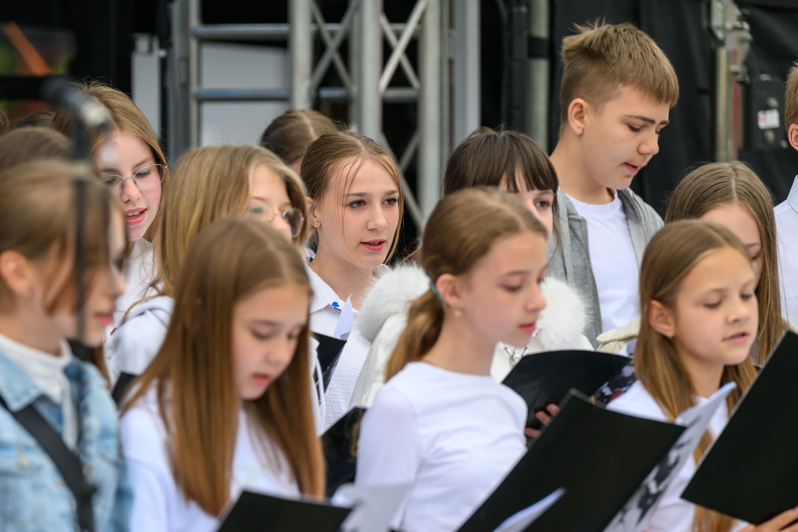 Grupa dzieci śpiewa w chórze na scenie, w białych strojach. Za nimi widać konstrukcję aluminiową sceny. Dzieci skupione na występie.