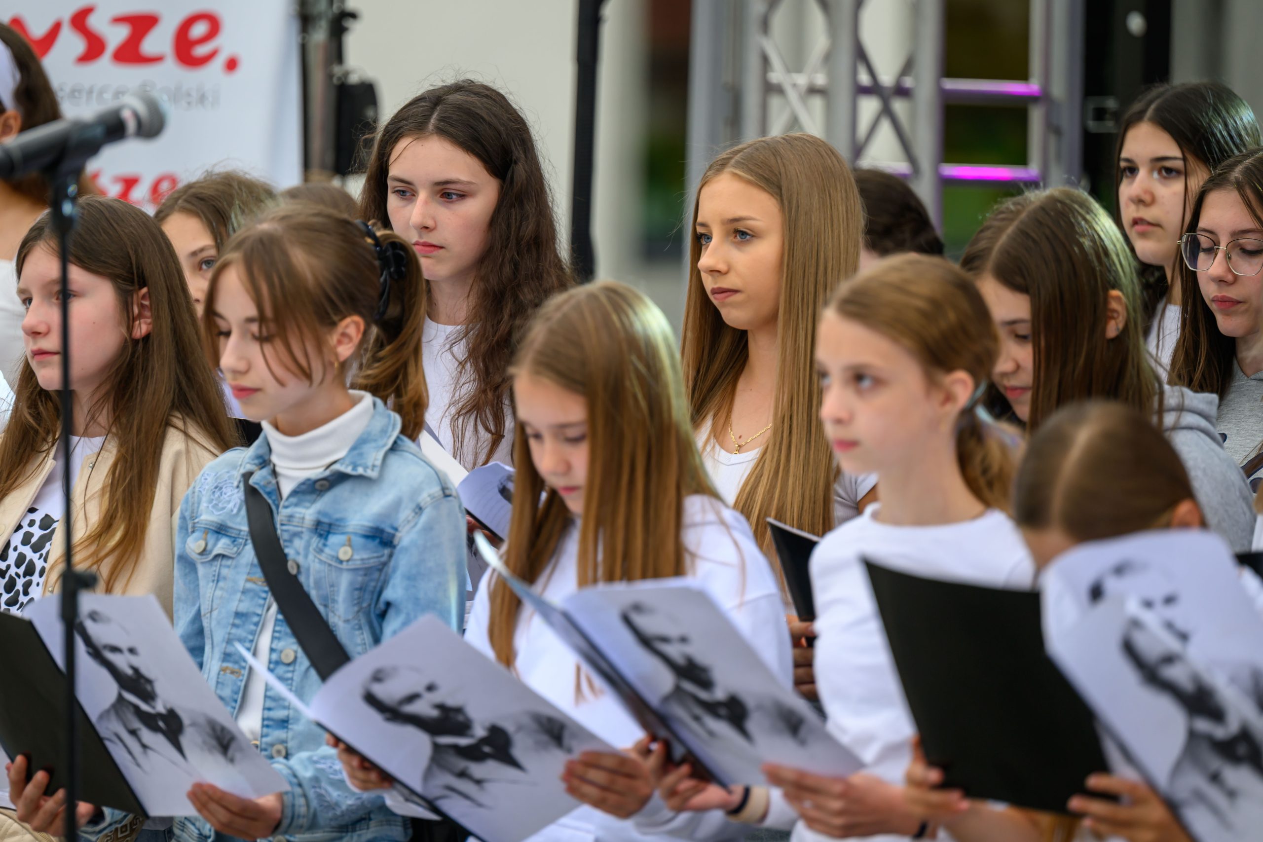 Grupa młodych dziewcząt w białych koszulkach stoi na scenie, trzymając kartki z czarno-białymi zdjęciami. W tle widoczny jest mikrofon i częściowo zarysowany napis. Kompozycja jest jasna i dynamiczna.