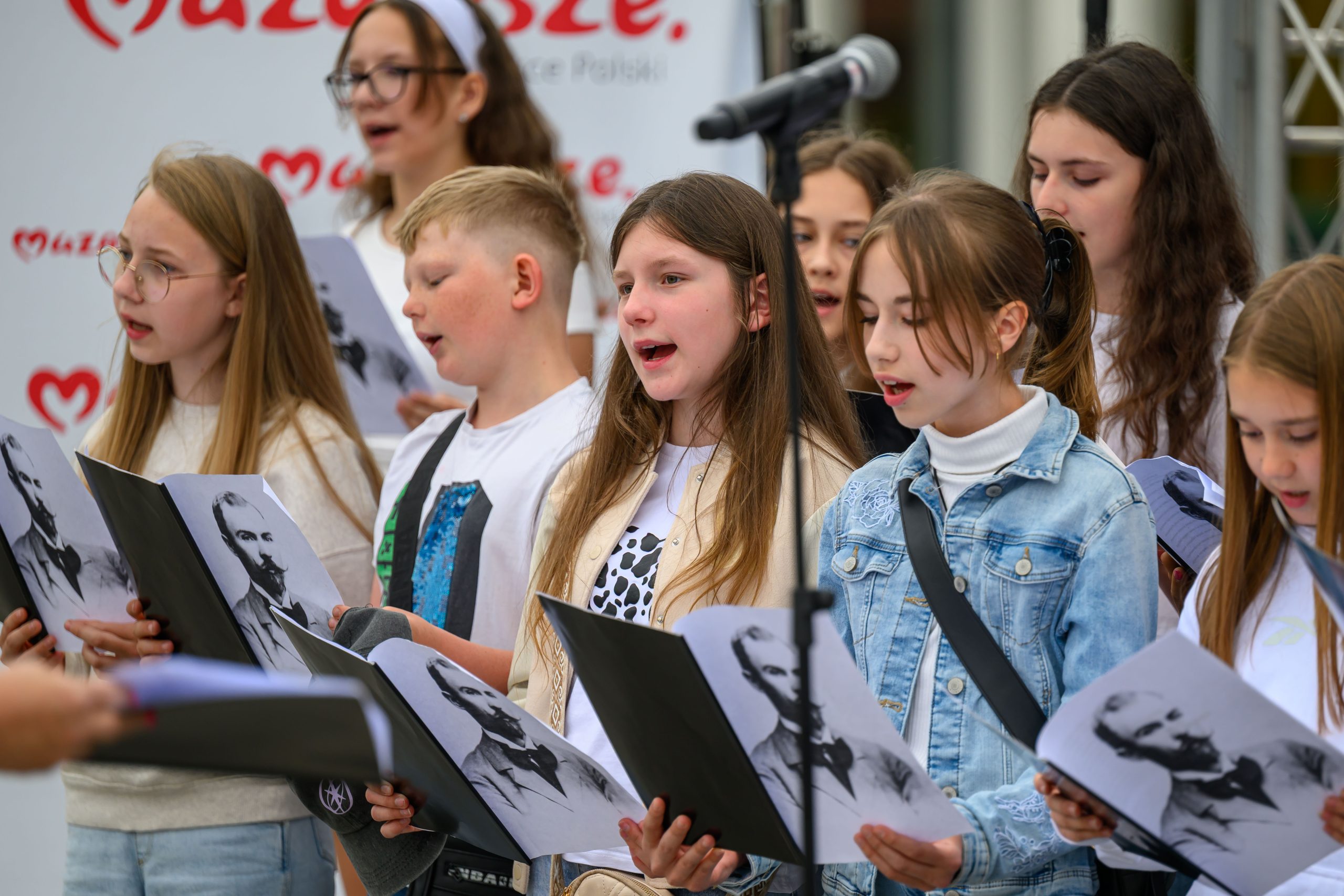Grupa dzieci śpiewa, trzymając nuty z czarno-białymi portretami. Są w jasnych ubraniach. W tle logo i mikrofon.