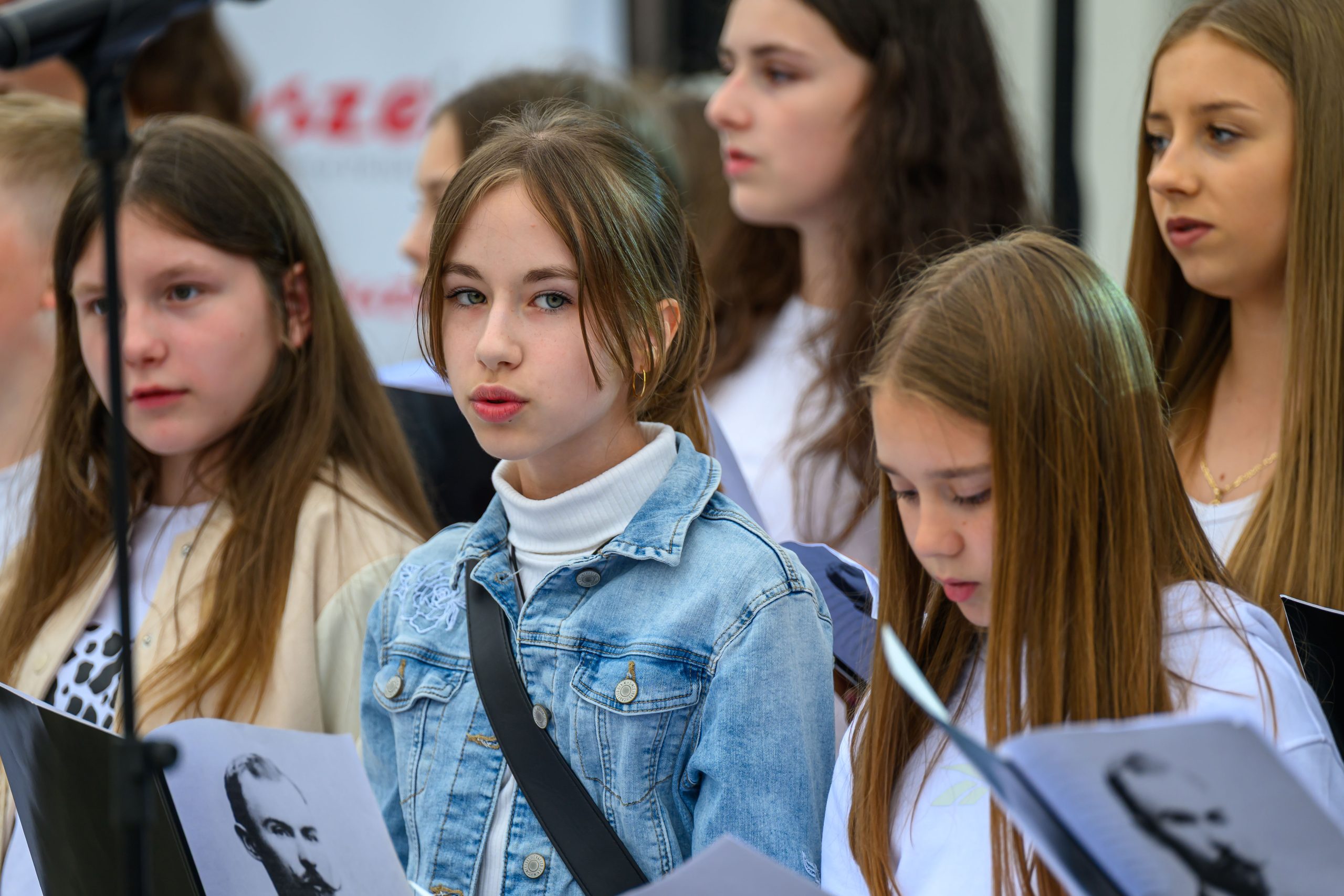 Grupa młodych osób, ubrana głównie na biało, śpiewa na scenie. Środkowa dziewczyna ma jeansową kurtkę i skupiony wyraz twarzy. W tle widać niewyraźne kształty i napis.