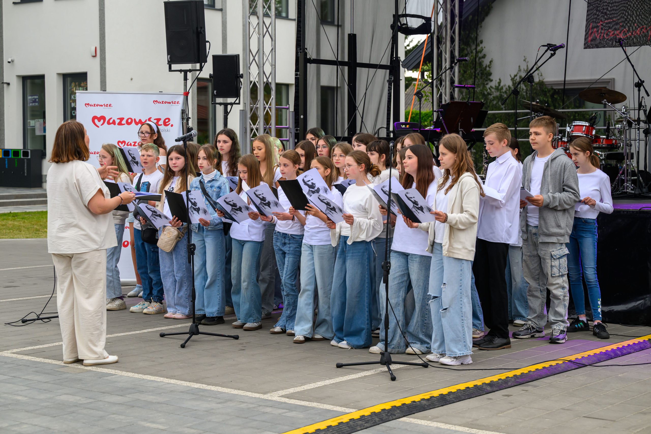 Grupa młodych osób śpiewa na scenie na świeżym powietrzu. Ubrani są w białe bluzki i dżinsy. W tle widoczne są instrumenty muzyczne i baner z napisem Mazowsze.