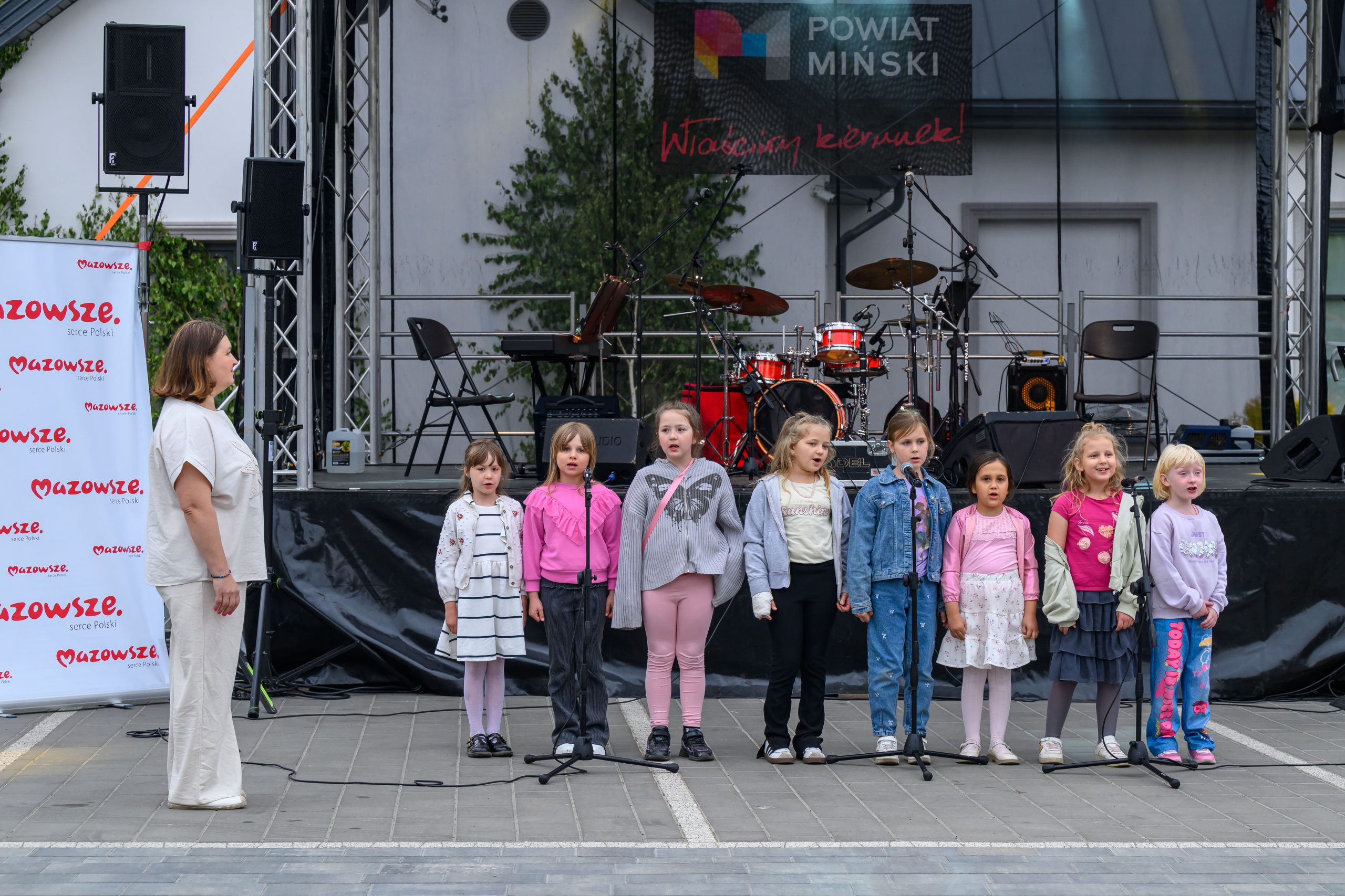 Na scenie dzieci śpiewają w kolorowych strojach. W tle instrumenty muzyczne i baner Mazowsze. Szare, zewnętrzne otoczenie.