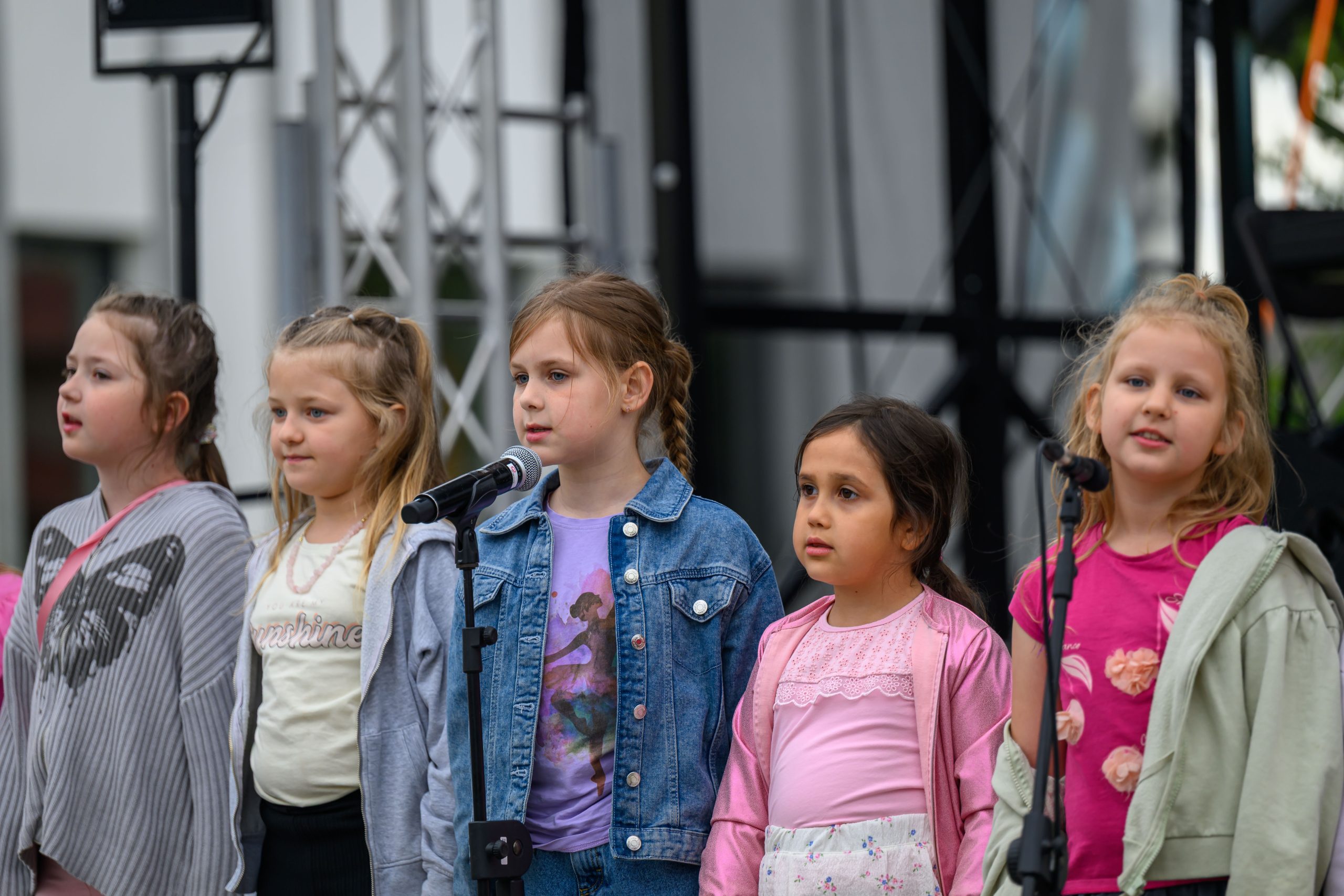 Na scenie pięć dziewczynek śpiewa przy mikrofonach. Mają kolorowe ubrania, m.in. różowe i niebieskie. W tle widać konstrukcję sceny.