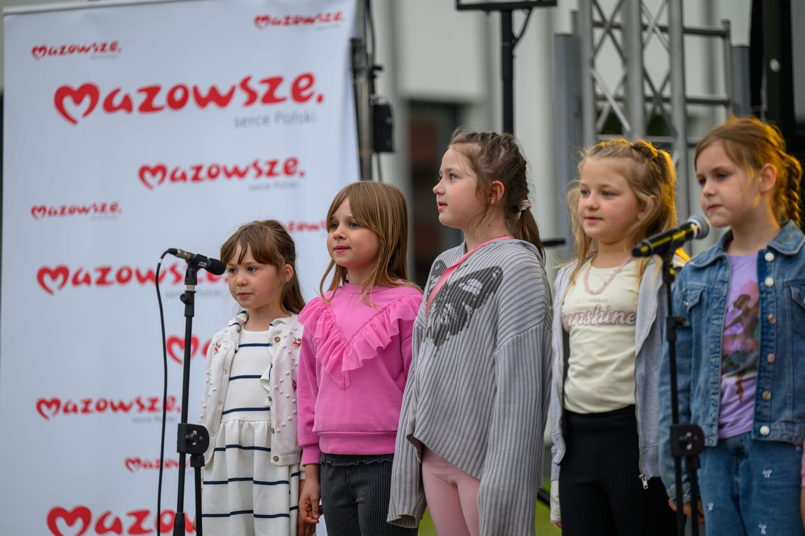 Pięć dziewczynek śpiewa na scenie na tle baneru z napisem Mazowsze. Ubrane są kolorowo, stoją przy mikrofonach. Tło jest jasne.