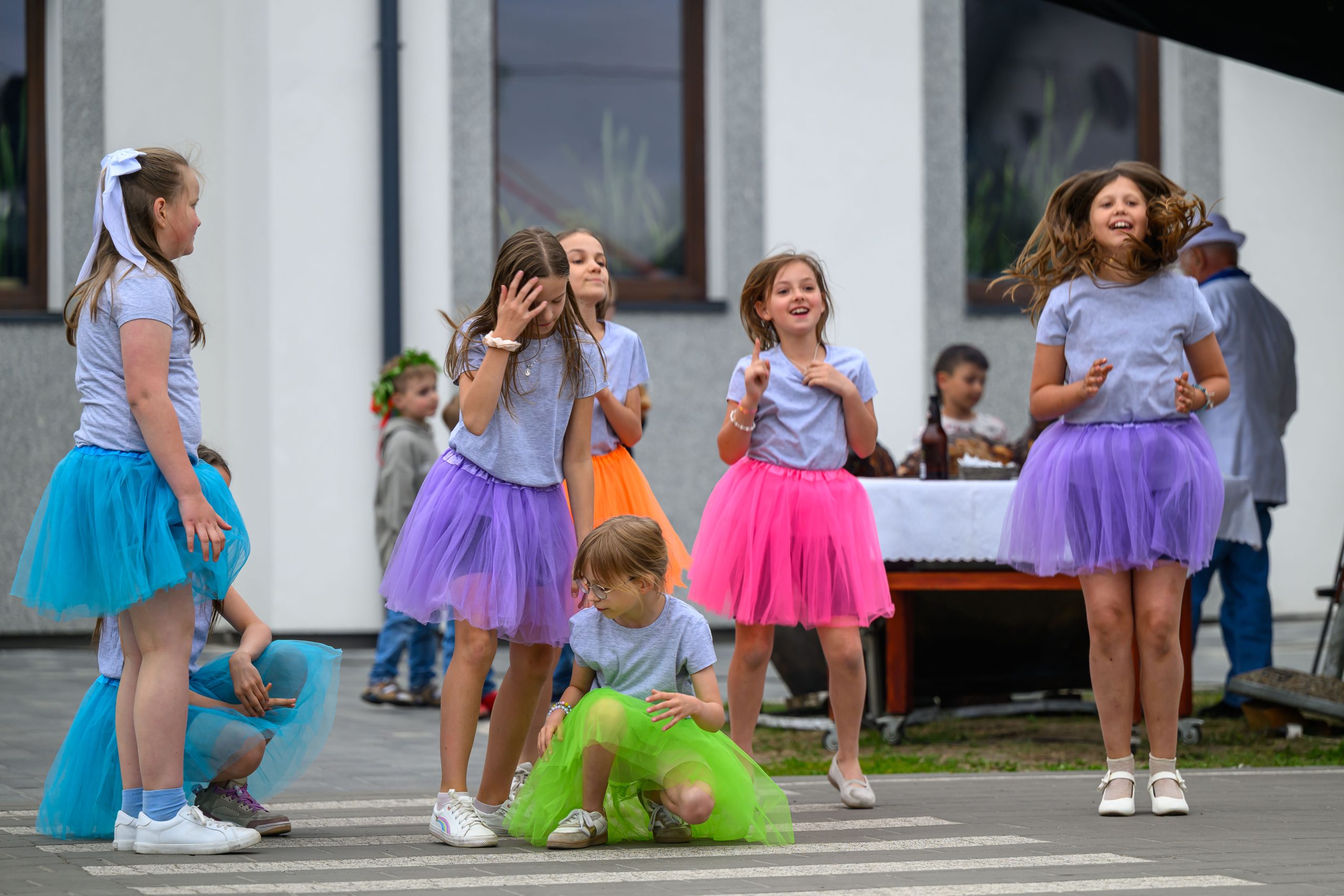 Grupa dziewczynek w kolorowych spódniczkach tańczy na zewnątrz. Tło to biały budynek z oknami. Ich stroje są jaskrawe i radosne.