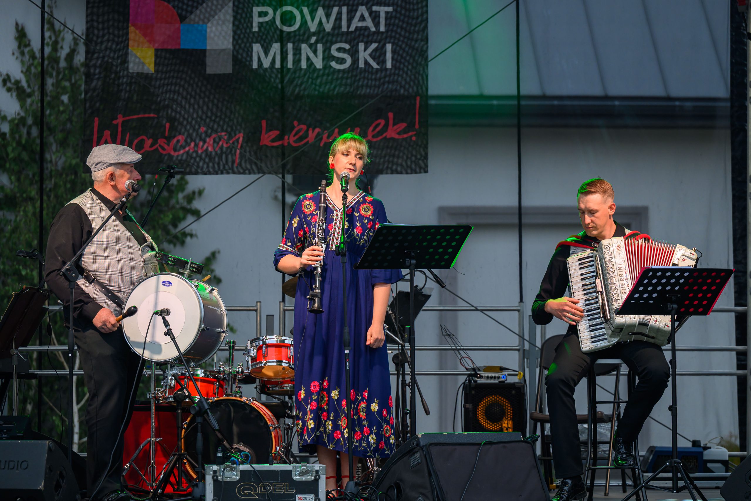 Trzy osoby grają muzykę na scenie. Kobieta w ludowej sukience gra na klarnecie, obok mężczyźni z bębnem i akordeonem. Tło z bębnem i zielenią.