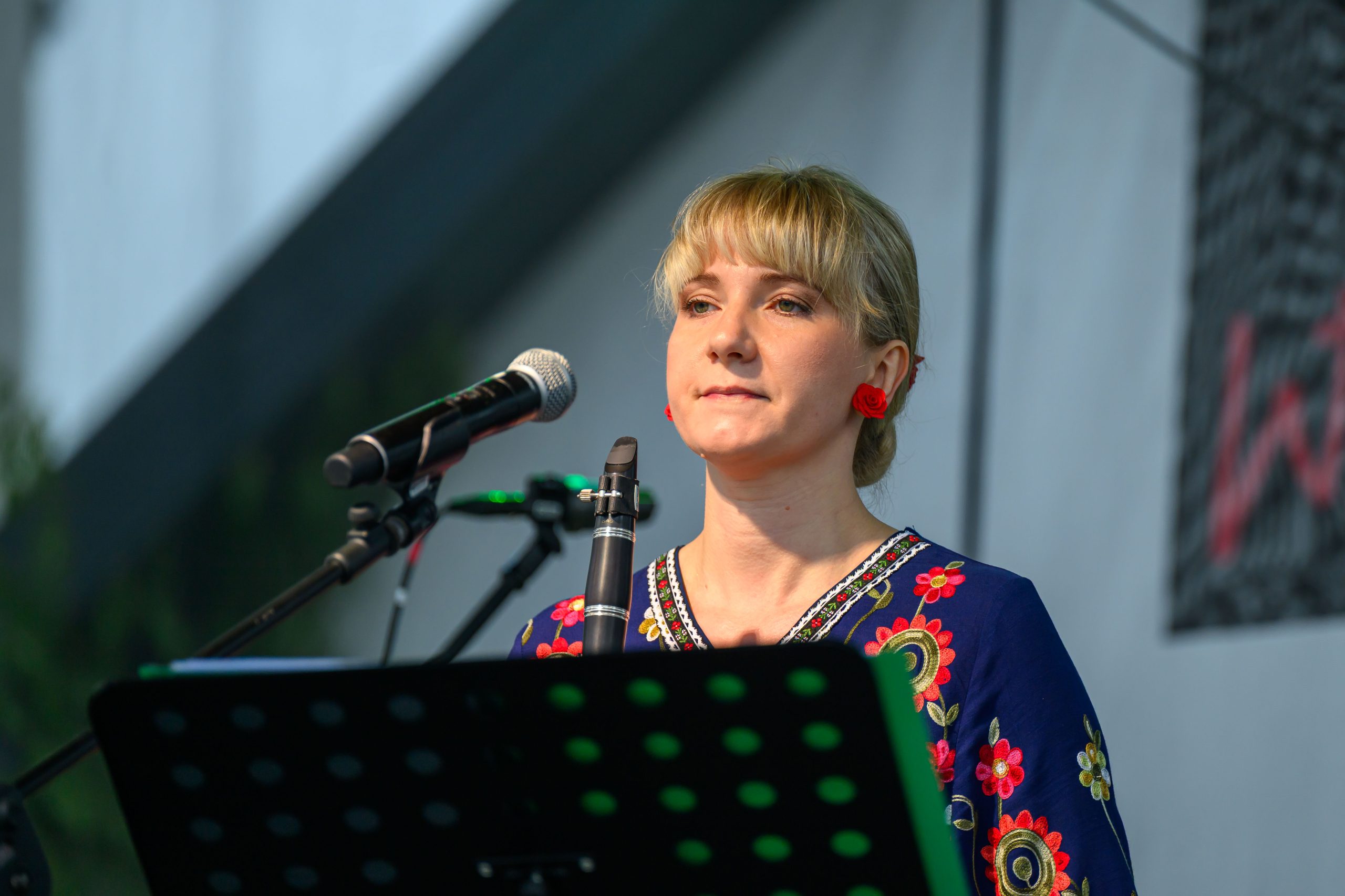 Kobieta na scenie w kolorowej sukni z haftem, trzyma klarnet przy mikrofonie. Tło szare, rozmyte. Widać nuty na stojaku.