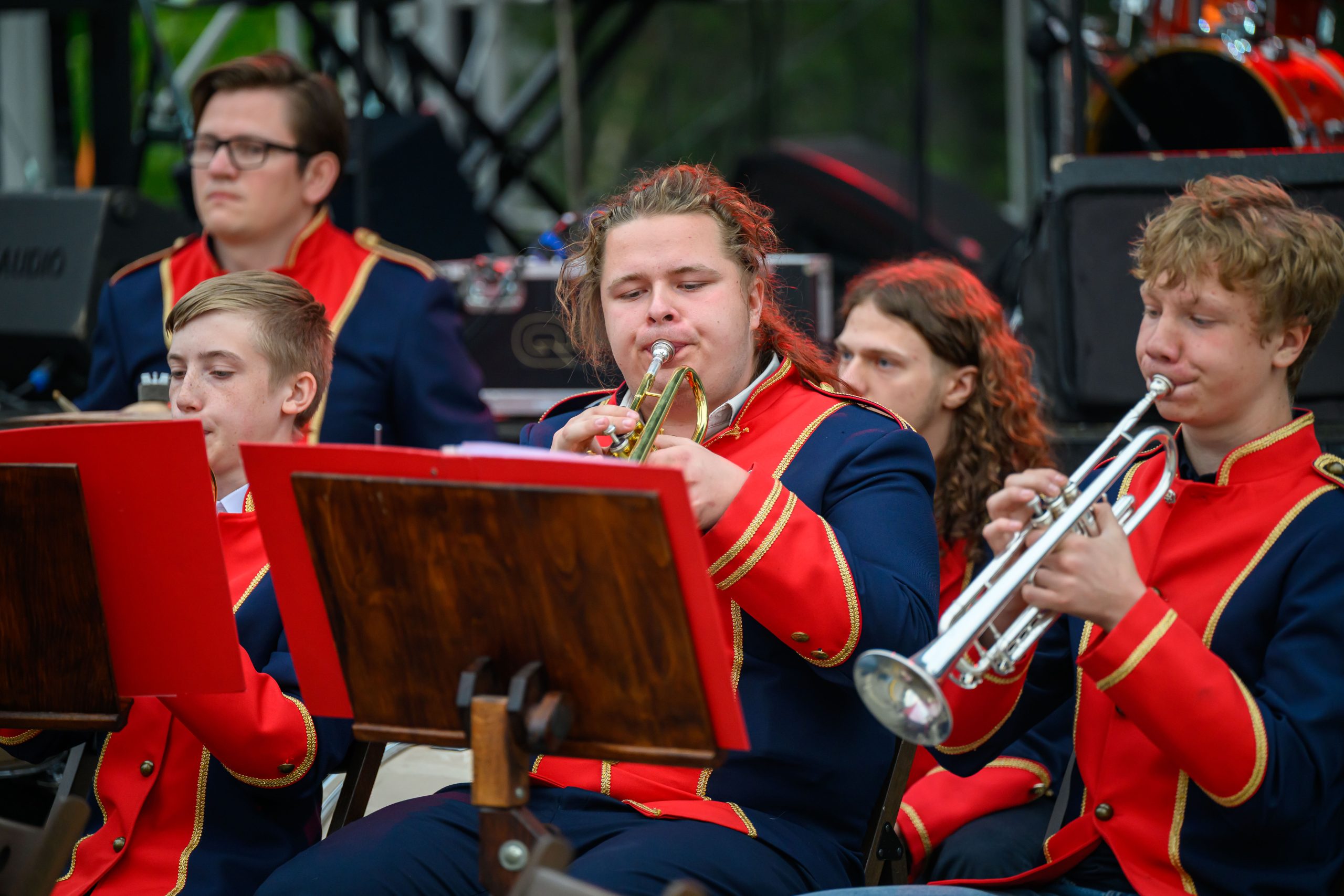 Grupa młodych muzyków w czerwono-granatowych uniformach gra na trąbkach. Tło ciemne, sceniczne, z elementami sprzętu muzycznego. Atmosfera koncertu na świeżym powietrzu.
