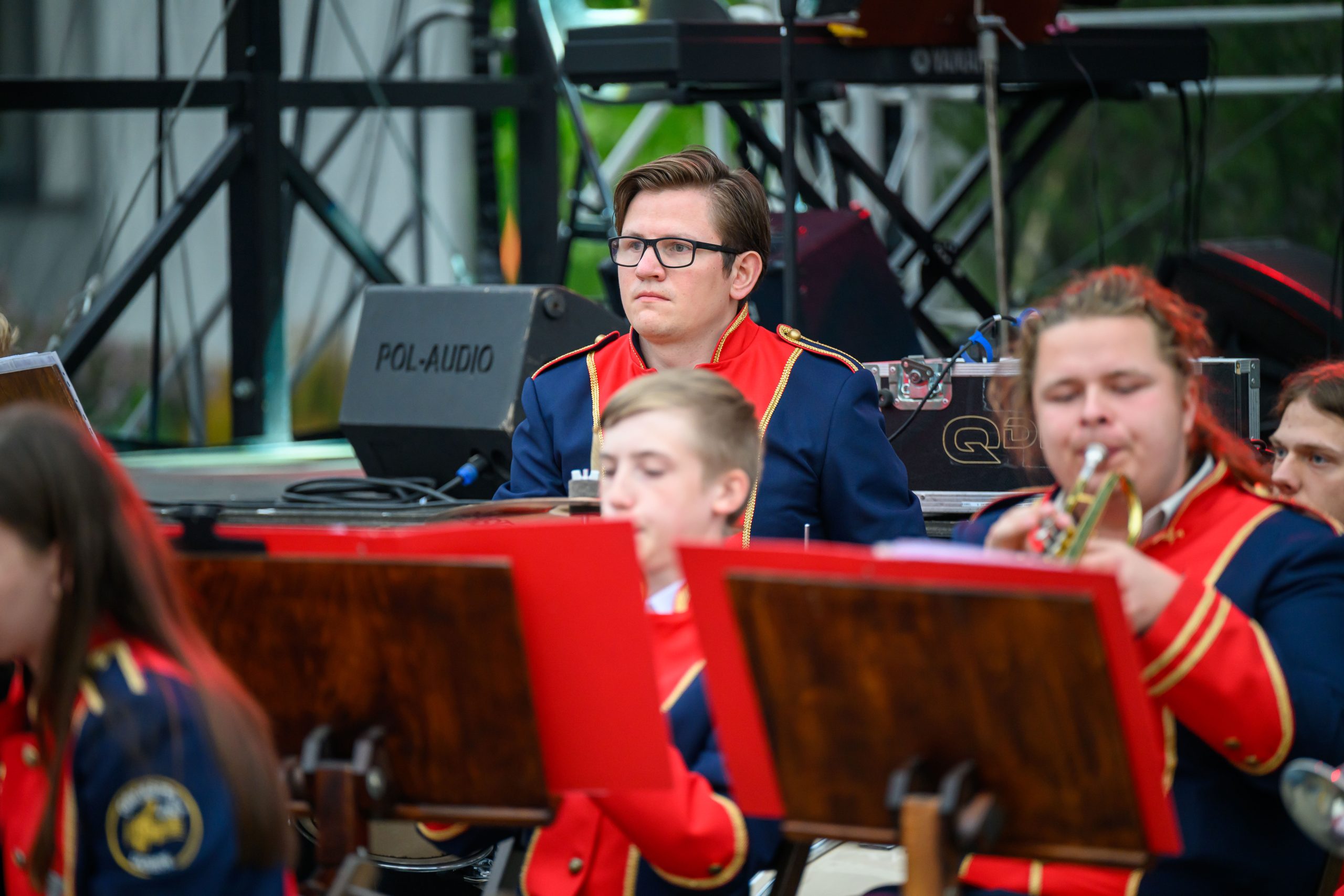 Grupa muzyków w granatowo-czerwonych mundurach na scenie. Mężczyzna z okularami siedzi z poważnym wyrazem twarzy. W tle sprzęt nagłaśniający.