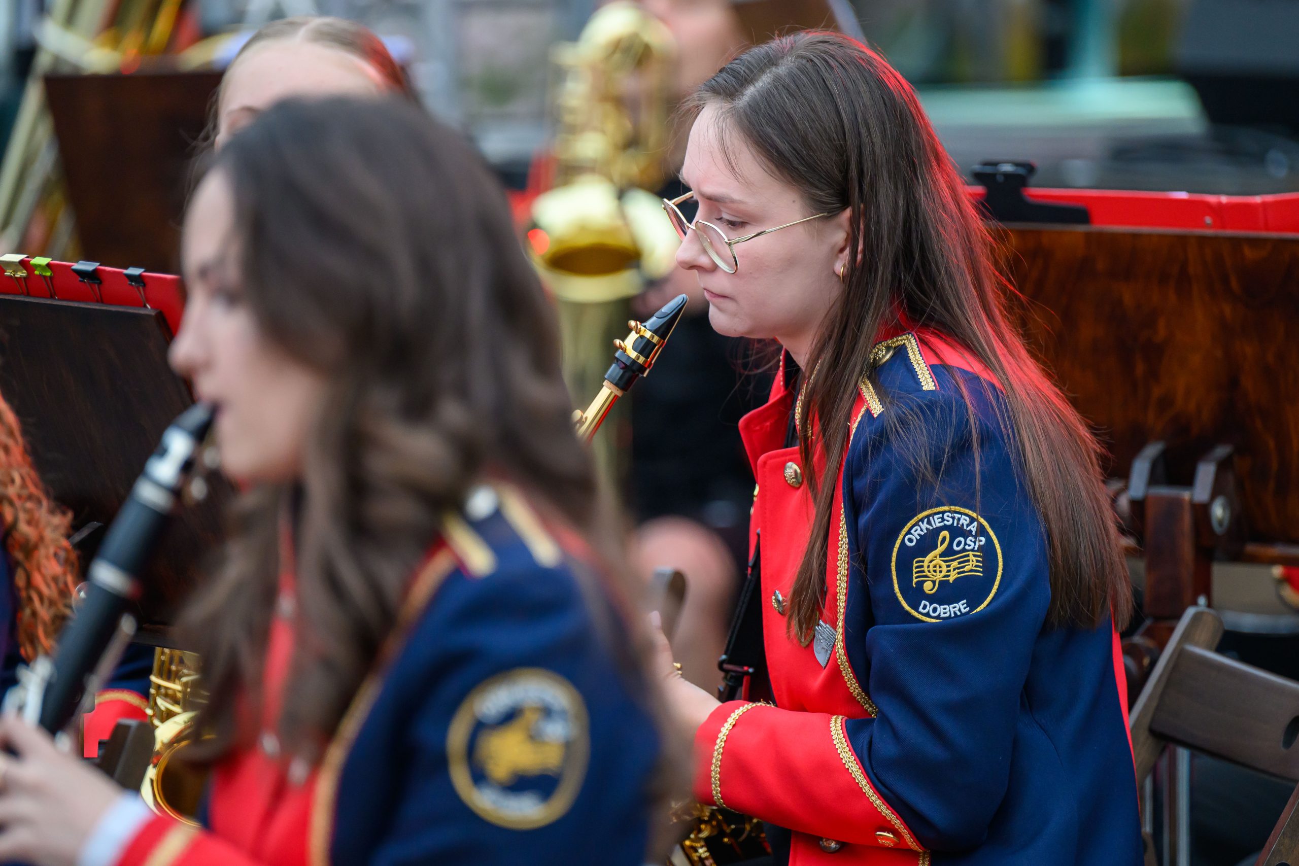Orkiestra dęta w kolorowych, granatowo-czerwonych mundurach. Muzycy grają na instrumentach dętych. Tło rozmyte.