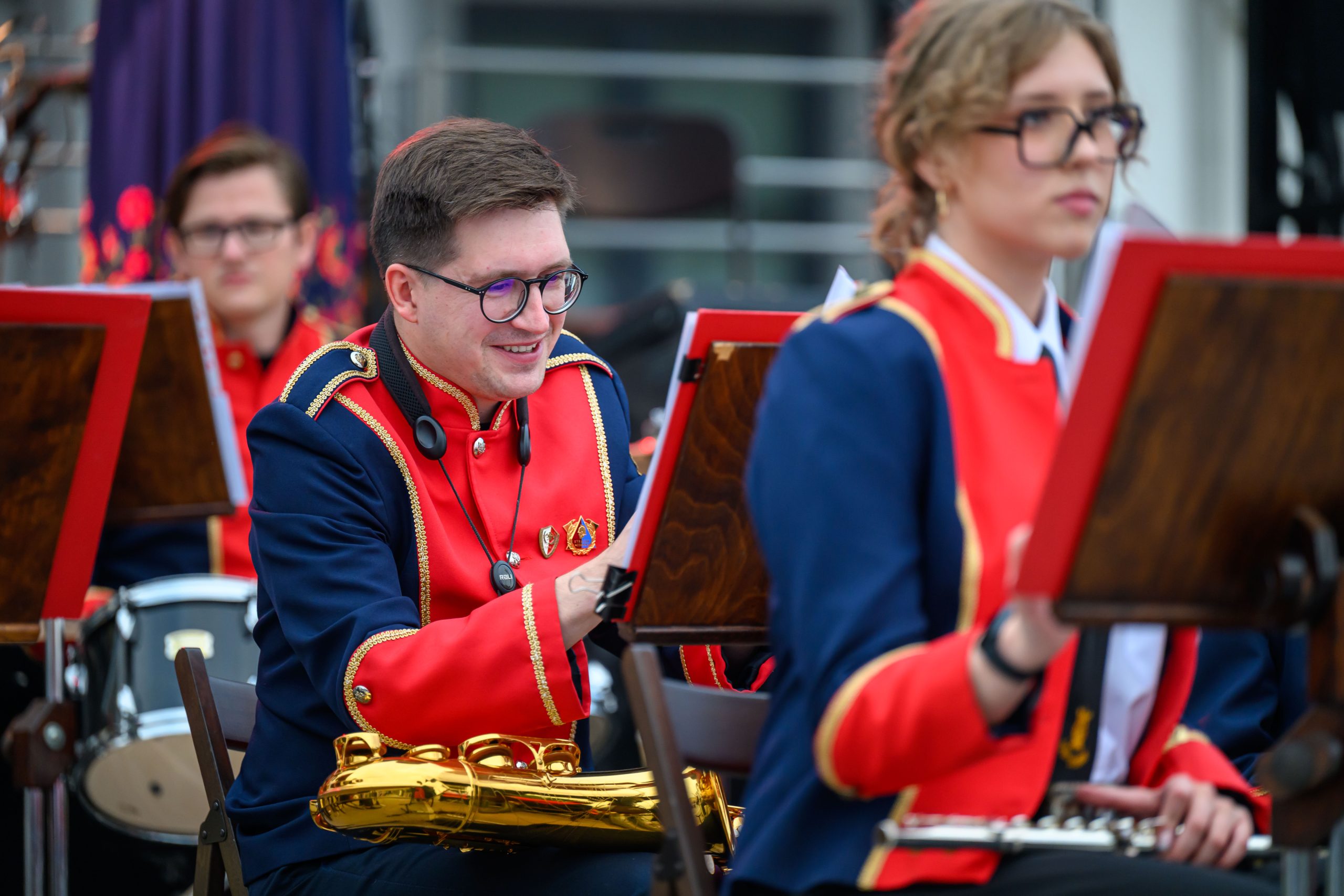Muzycy w orkiestrze w czerwono-granatowych mundurach grają na instrumentach. Na pierwszym planie saksofon. Tło jest rozmyte.
