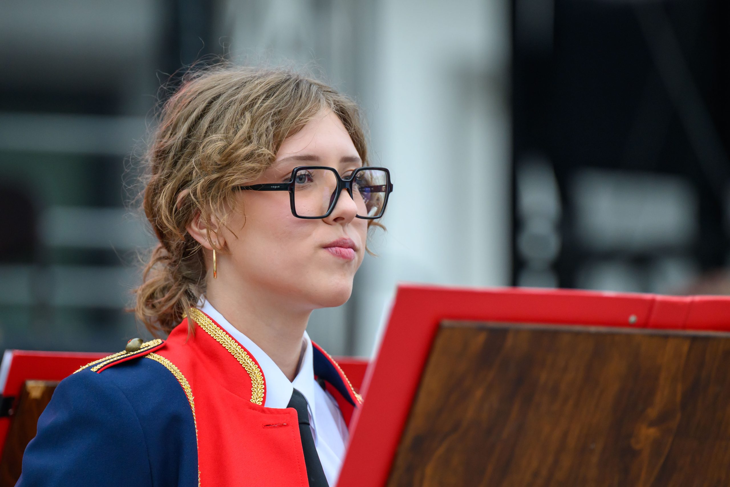 Osoba w okularach i mundurze orkiestry marszowej, z czerwonym szalikiem. W tle rozmyte szczegóły budynku. Kolory to głównie czerwień i granat.