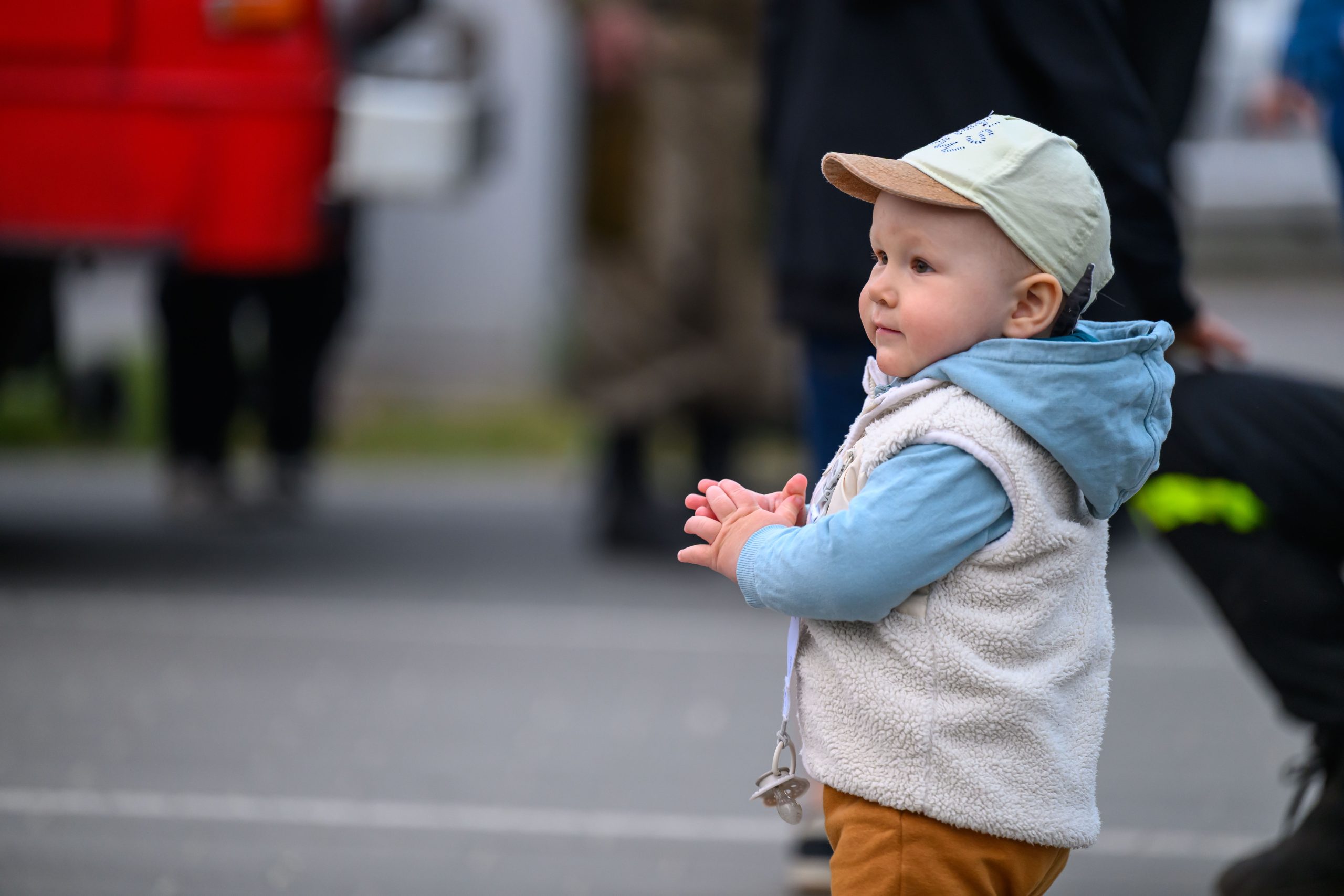 Małe dziecko w czapce i niebieskiej bluzie stoi na zewnątrz. Tło jest rozmyte, widać czerwony kształt. Dziecko ma zawieszkę na smoczku.