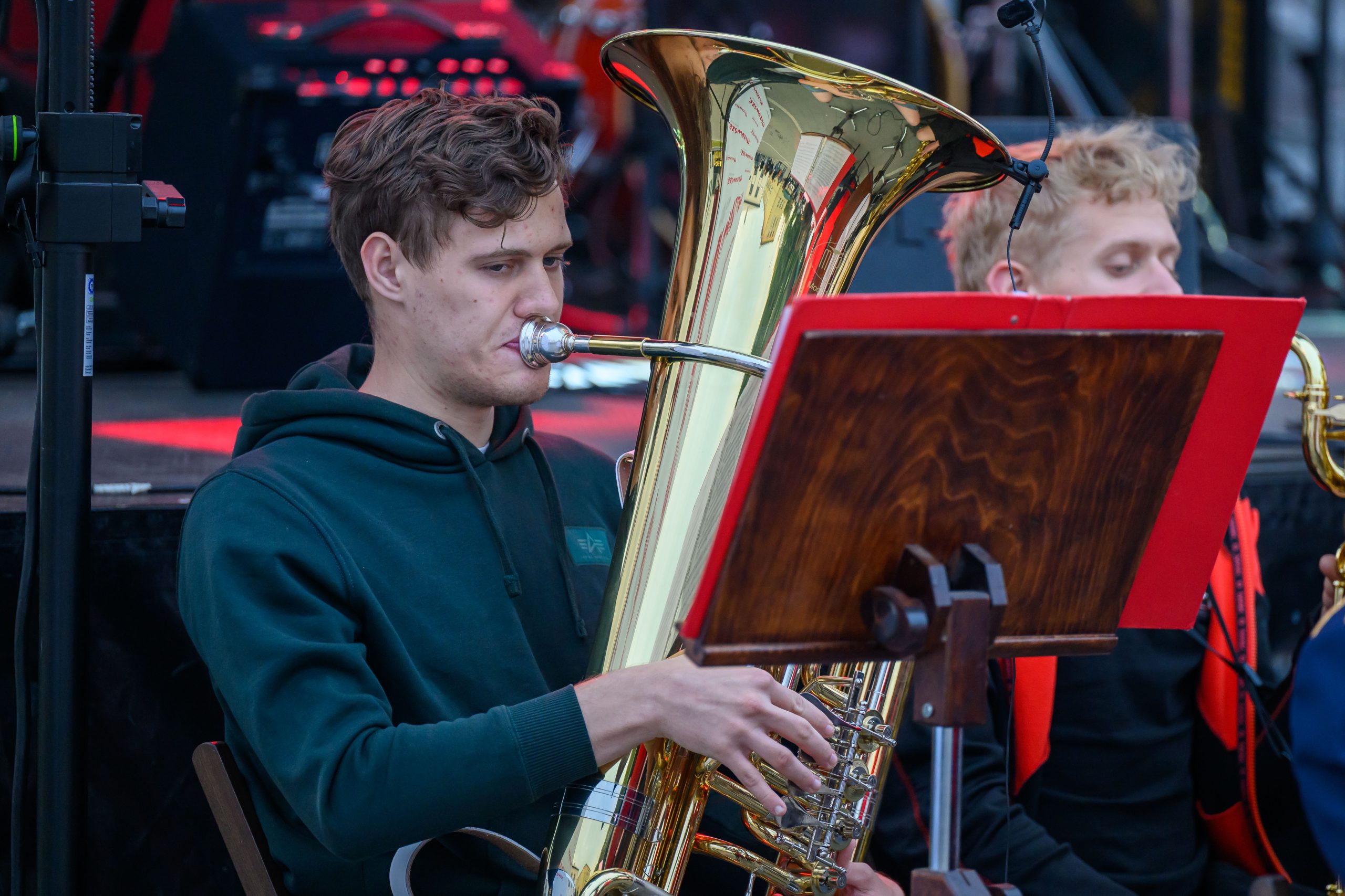 Muzyk gra na tubie podczas koncertu. Instrument mieni się złotym kolorem, a w tle widać sceniczne oświetlenie. Młody mężczyzna nosi zieloną bluzę, a przed nim znajduje się pulpit z nutami.