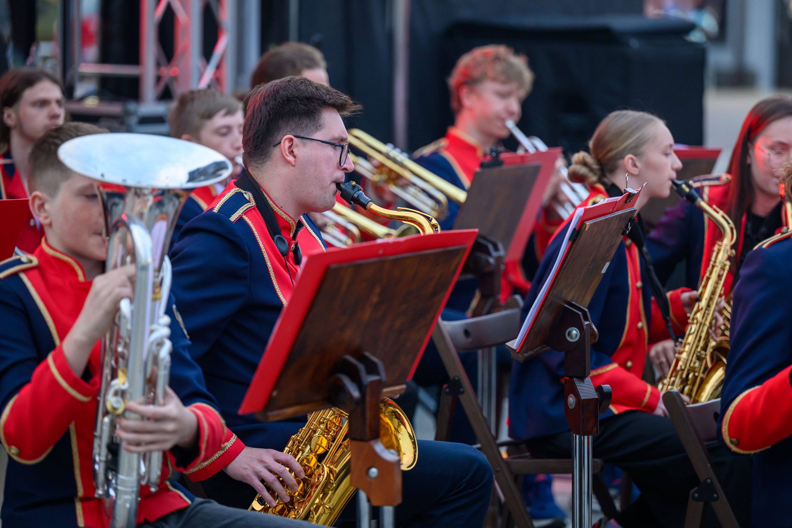 Orkiestra dęta w kolorowych, granatowo-czerwonych uniformach. Muzycy grają na instrumentach, głównie saksofonach. Tło rozmyte, sceniczne oświetlenie.