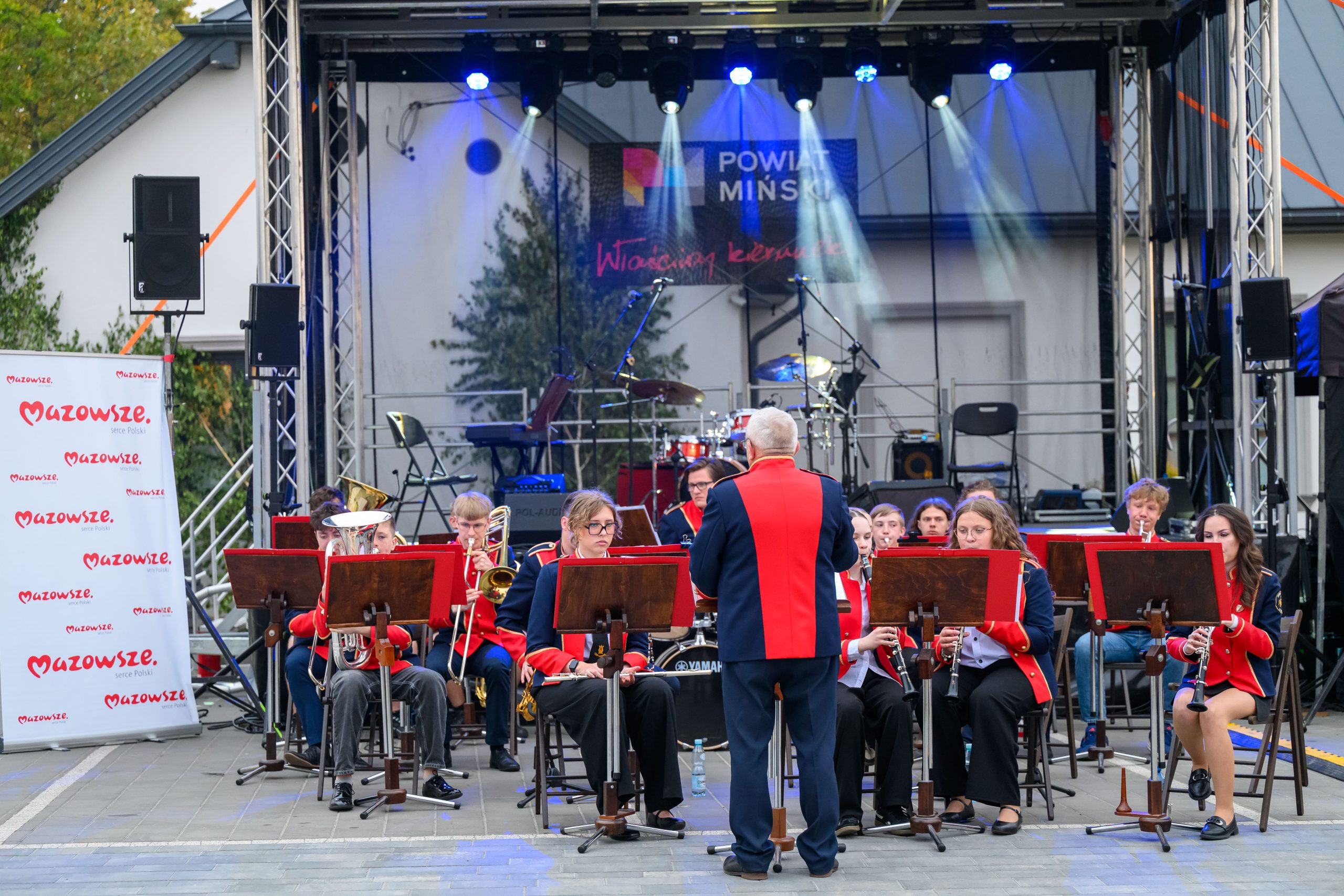 Orkiestra w czerwono-niebieskich mundurach gra na scenie. W tle wiszą reflektory i bębny. Scena ustawiona na zewnątrz.