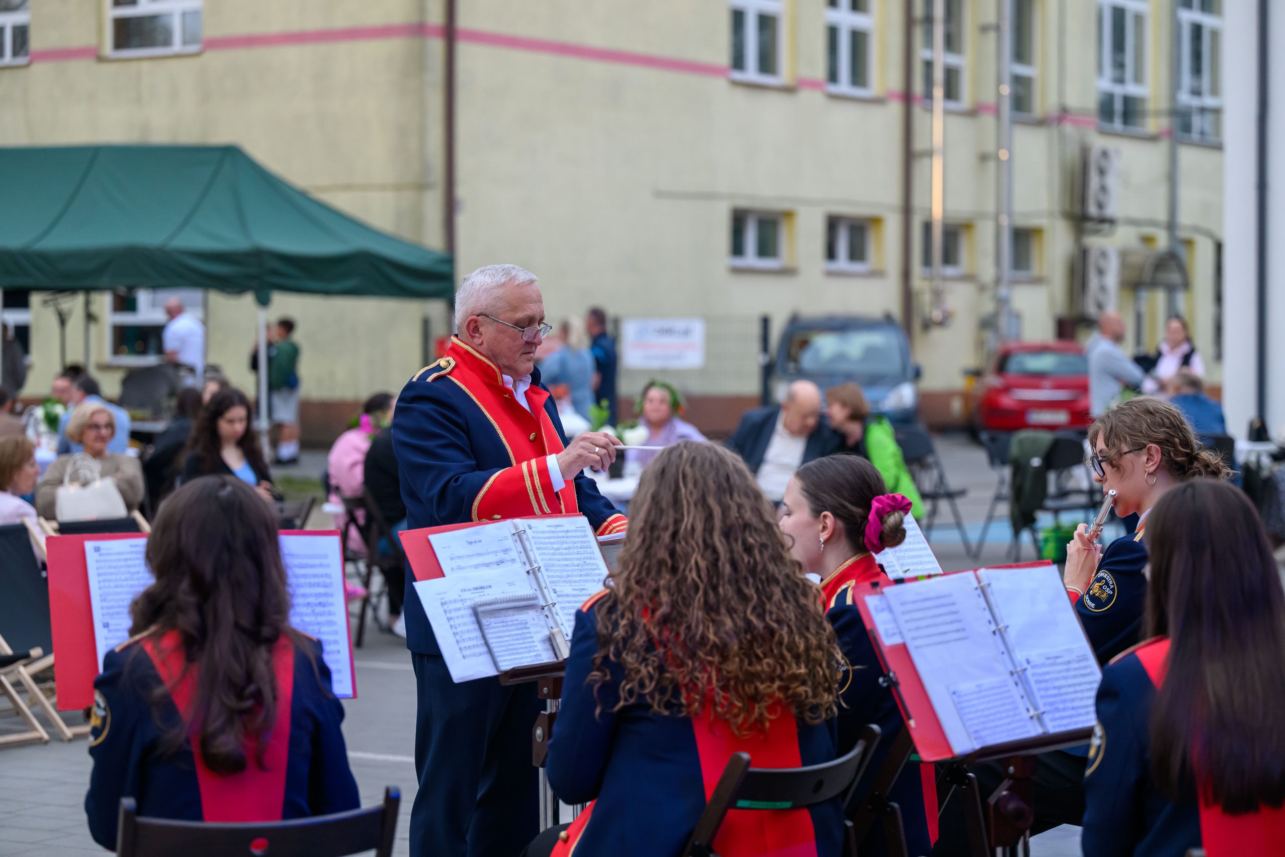Mężczyzna dyryguje orkiestrą w mundurach z czerwonymi elementami. W tle ludzie siedzą przy stolikach na zewnątrz budynku. Tło jest beżowe z zielonym namiotem.