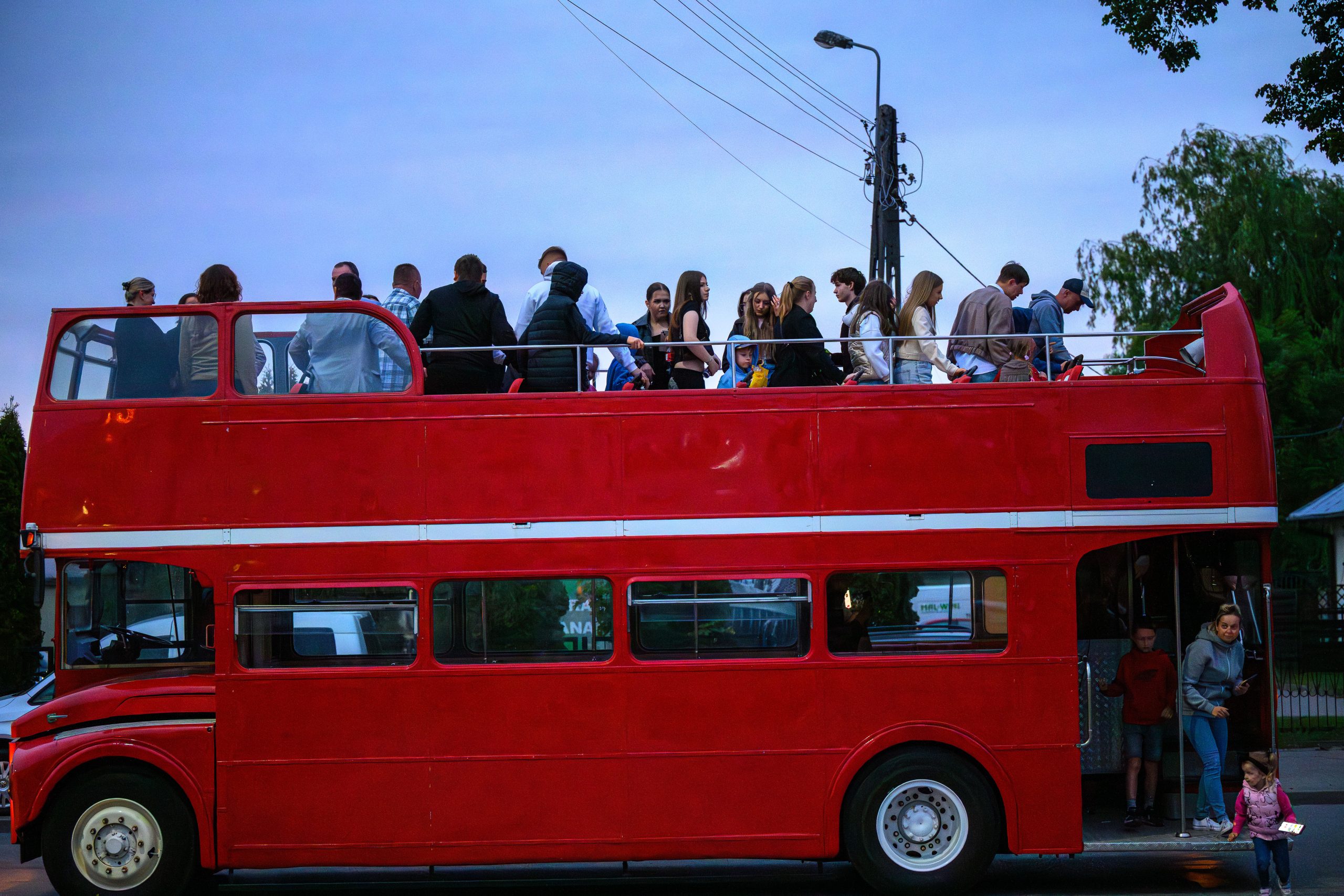 Czerwony piętrowy autobus z otwartym górnym pokładem wypełniony ludźmi. Wieczorne niebo, latarnia i drzewa w tle. W dolnej części kilka osób wsiada lub wysiada.
