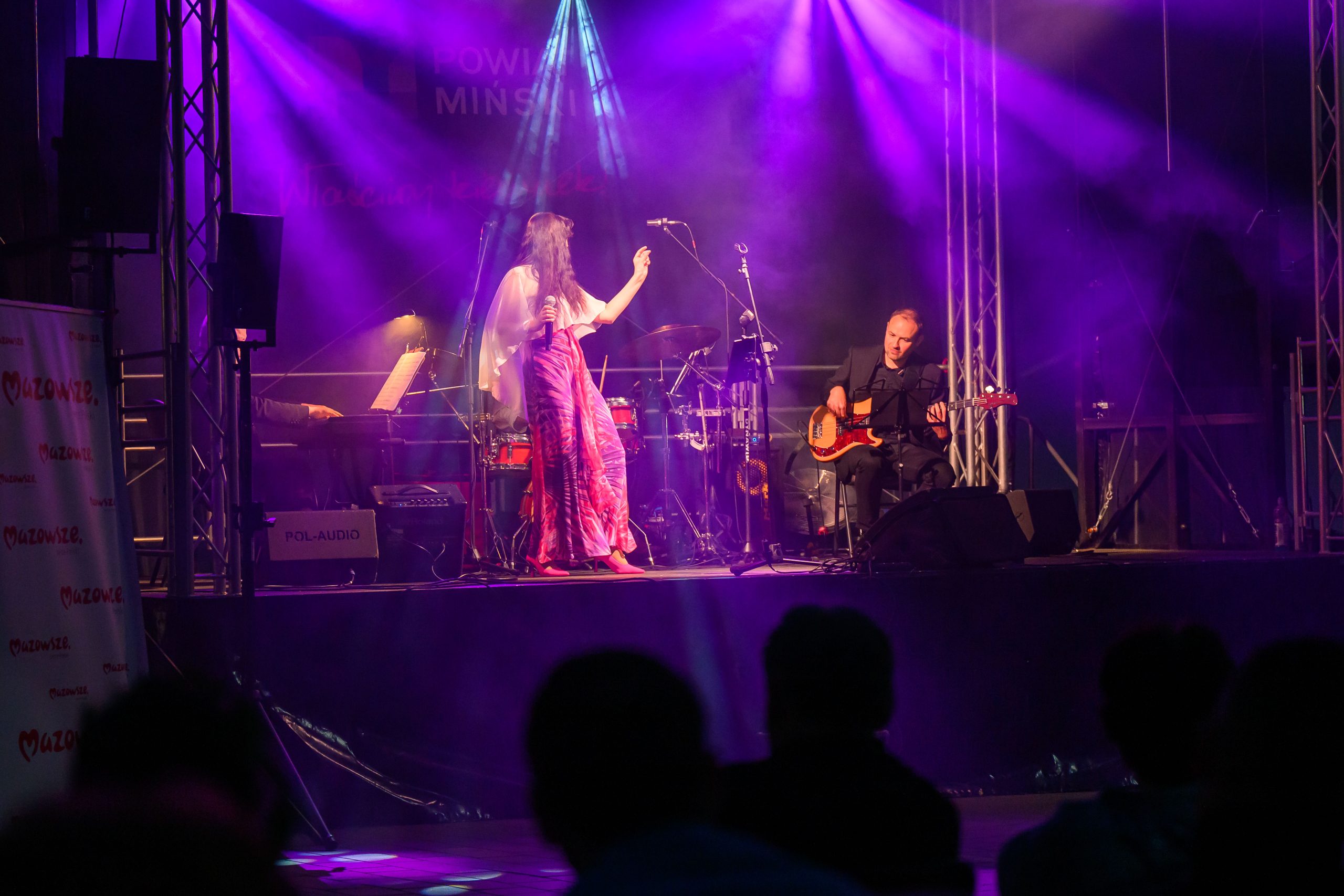 Na scenie oświetlonej fioletowymi światłami występuje wokalistka i gitarzysta. Kobieta ma długą suknię, a mężczyzna siedzi z gitarą. W tle perkusja i pianino, widoczni są także widzowie.