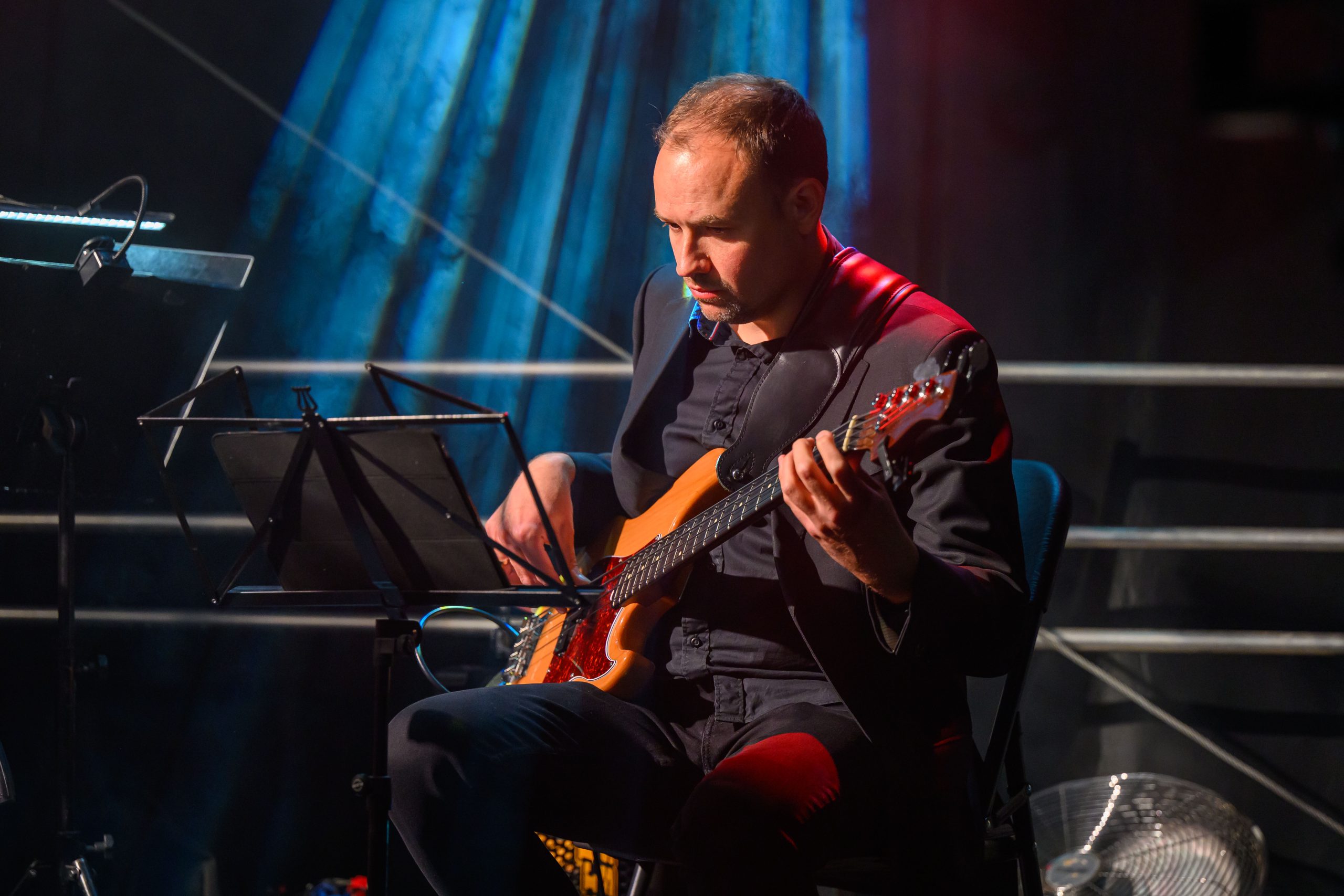 Muzyk gra na gitarze basowej w ciemnym ubraniu. Tło jest niebieskie z jasnym oświetleniem. Obok stojak na nuty.