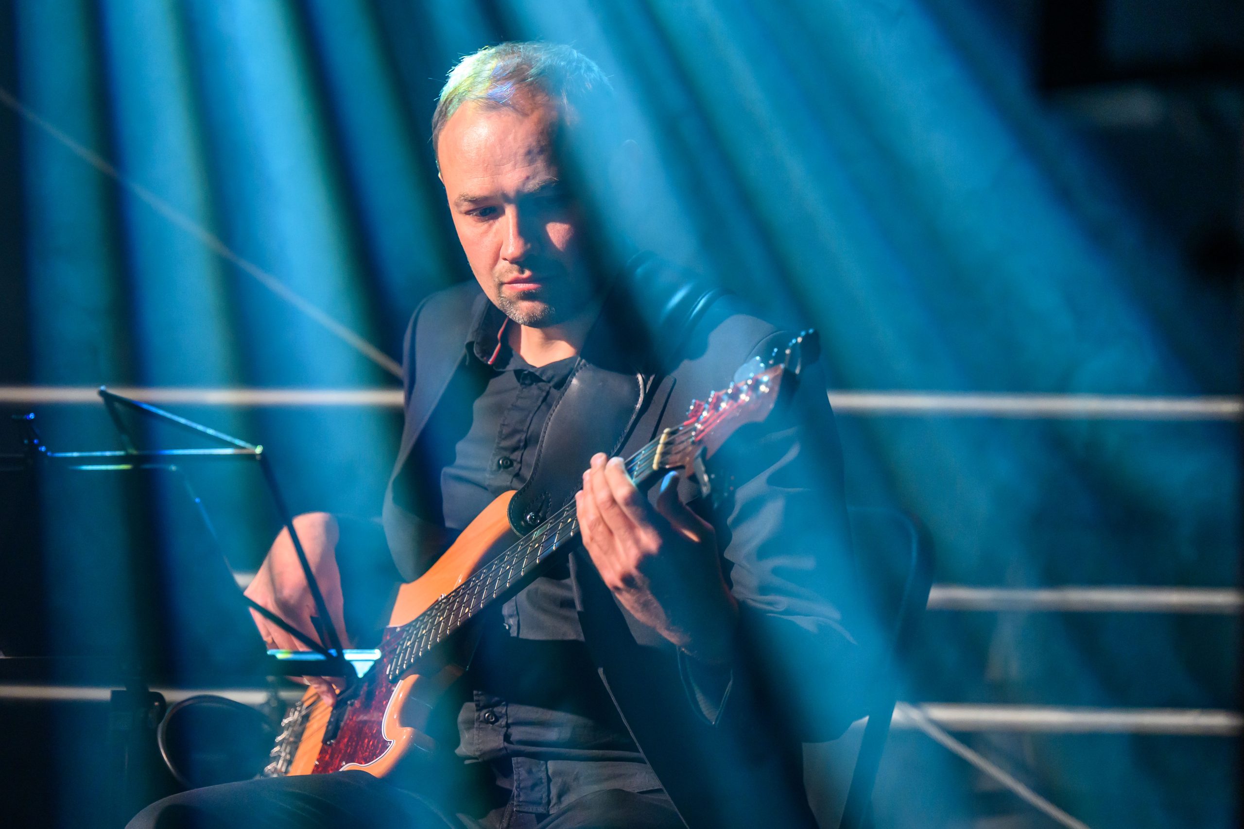 Mężczyzna gra na gitarze elektrycznej na scenie. Oświetlenie w niebieskich odcieniach. Tło ciemne z metalowymi barierkami.