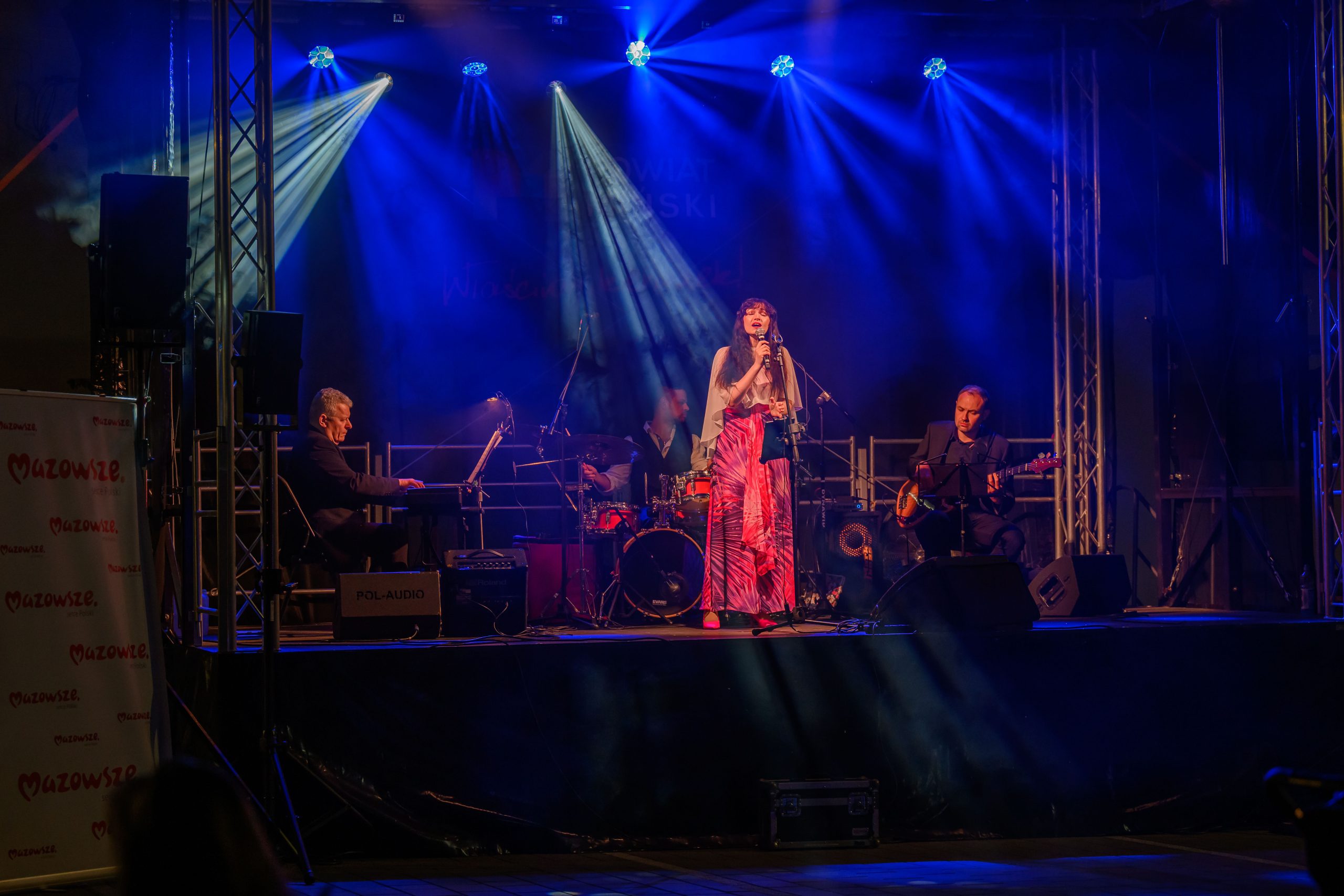 Koncert na scenie, wokalistka na środku w różowej sukni, muzycy po bokach. Oświetlenie niebieskie i fioletowe. Tło ciemne.