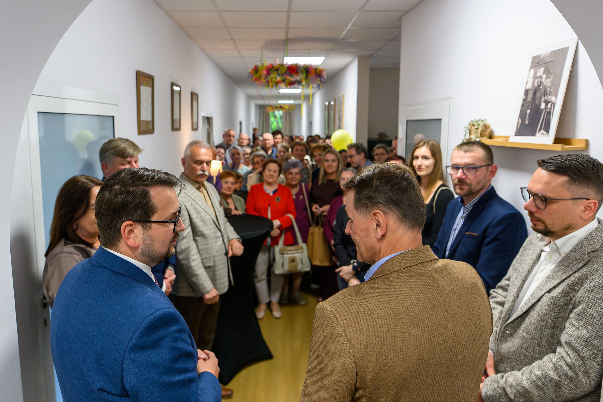 Grupa ludzi w korytarzu, większość w eleganckich strojach. Ściany białe, na nich obrazy. Na suficie kolorowe dekoracje.