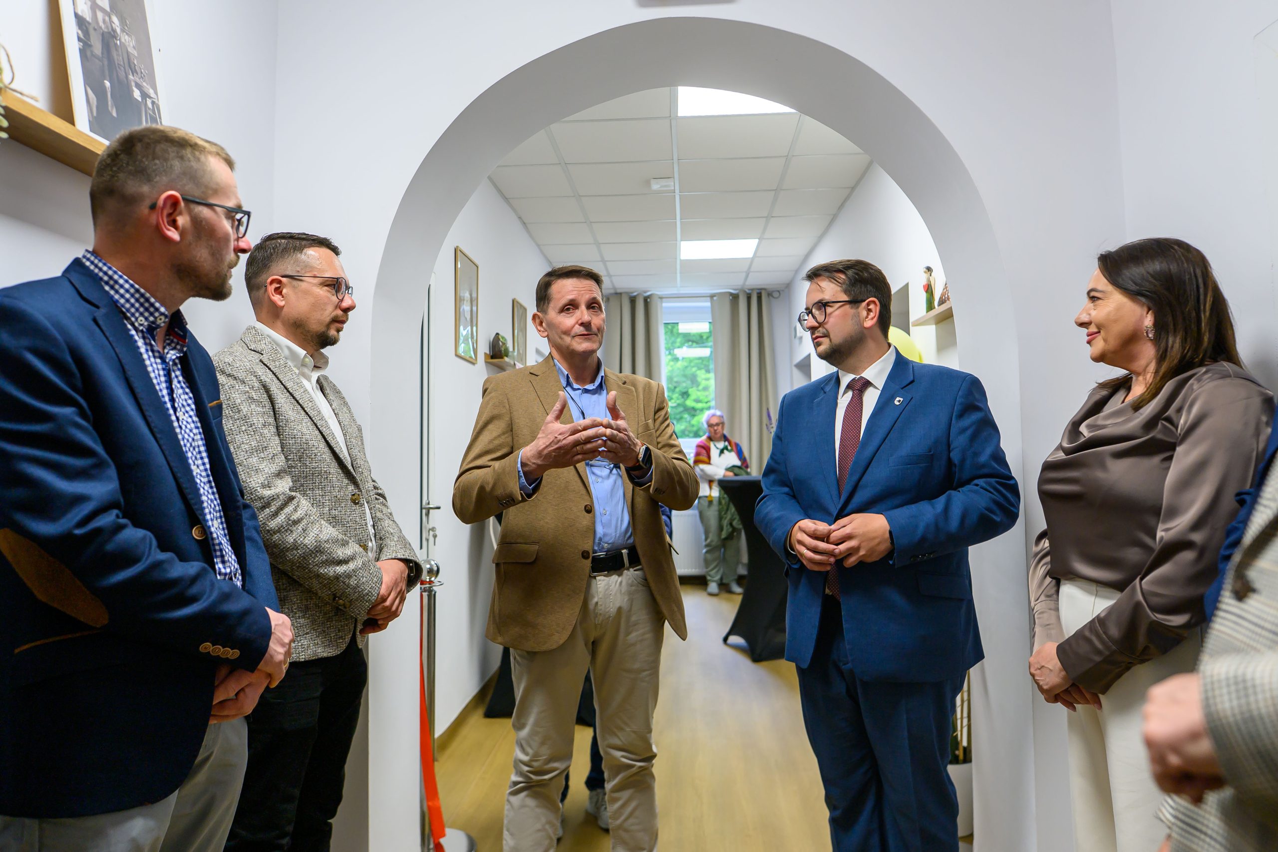 Sześć osób w korytarzu, eleganckie stroje, dyskutują. Ściany jasne, zdjęcia na półkach. Podłoga drewniana, okno w tle.