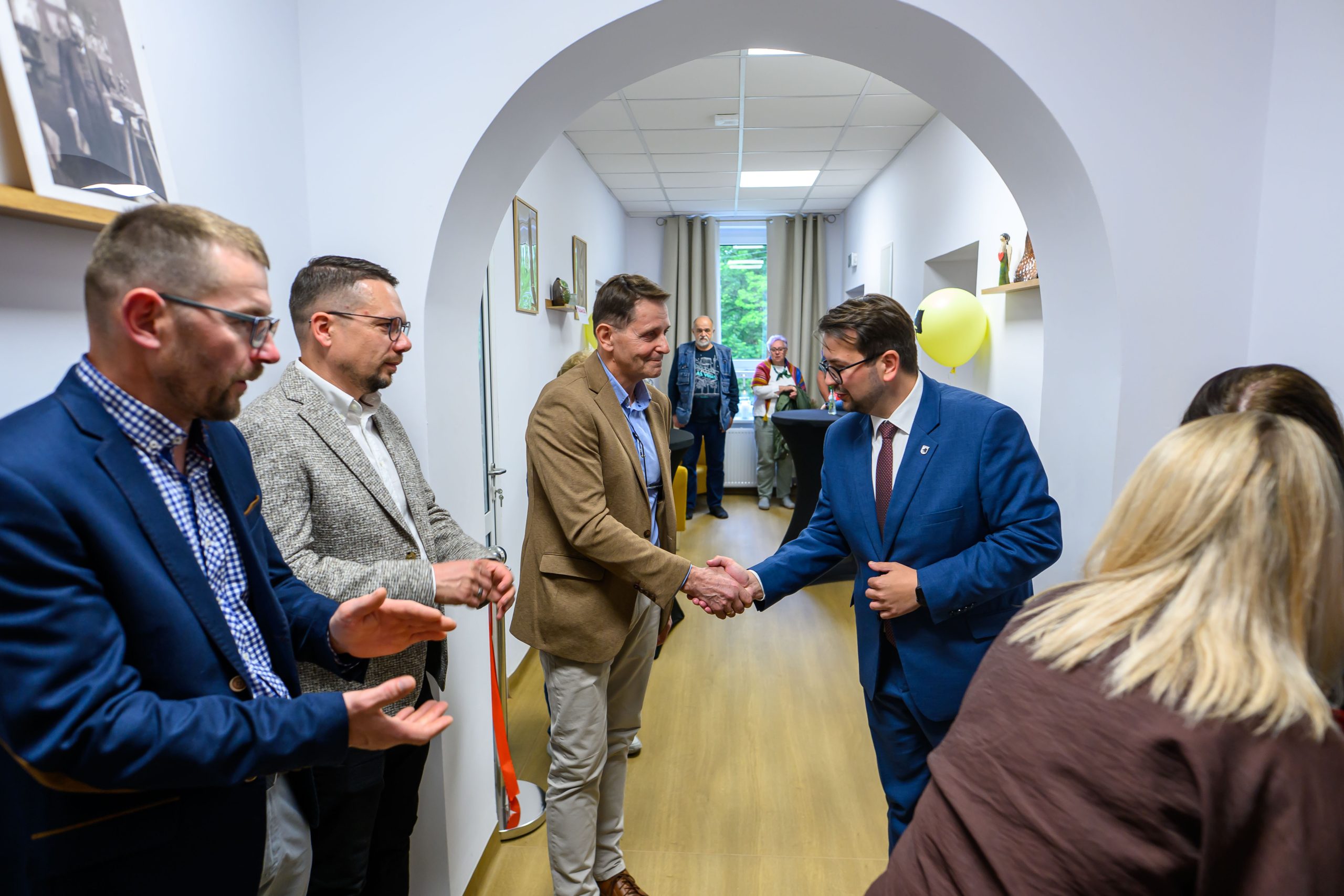 Grupa mężczyzn w garniturach wita się uściskiem dłoni w jasnym korytarzu. Tło to białe ściany z obrazami. Klimat formalny, wydarzenie w pomieszczeniu.