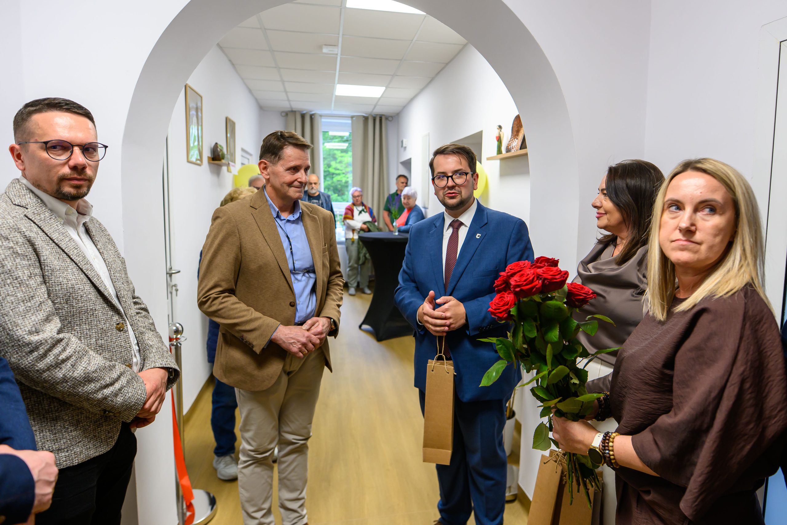 Grupa osób stoi w wąskim korytarzu, na pierwszym planie kobieta trzyma bukiet czerwonych róż. Ściany są białe, na podłodze drewniany panel. W tle widać okno i kilka obrazów na ścianach.