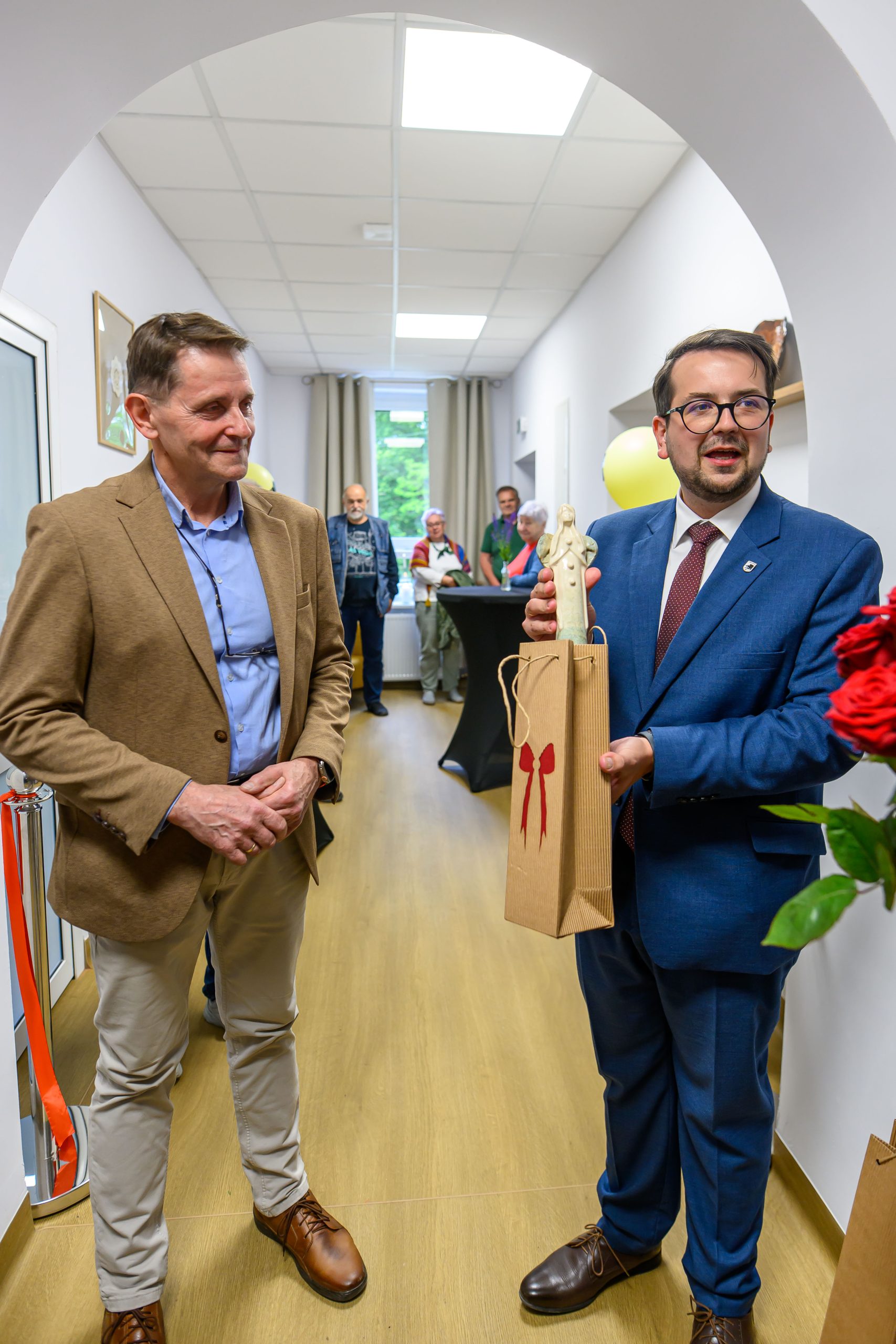 Dwóch mężczyzn stoi w korytarzu. Jeden trzyma torebkę z figurką. Ściany białe, podłoga drewniana, w tle kilka osób.