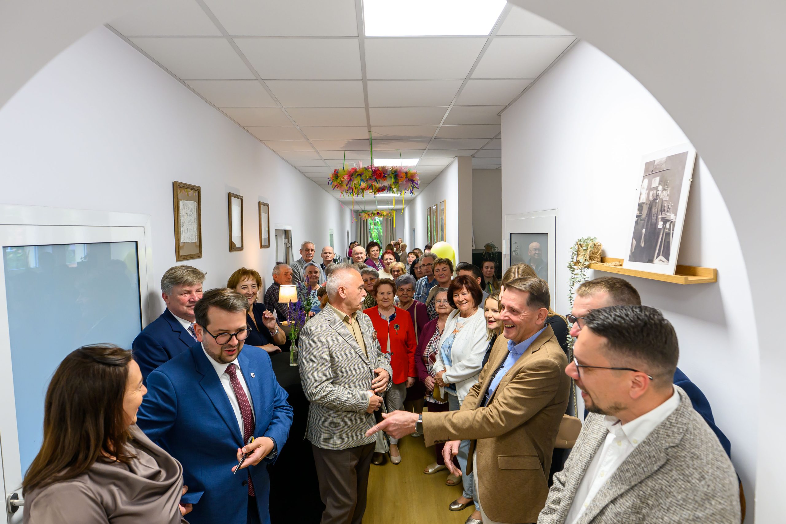 Grupa ludzi stoi w jasnym korytarzu, uśmiechnięci i rozmowni. W tle wiszą kolorowe dekoracje sufitowe. Ściany zdobią obrazy i fotografie.
