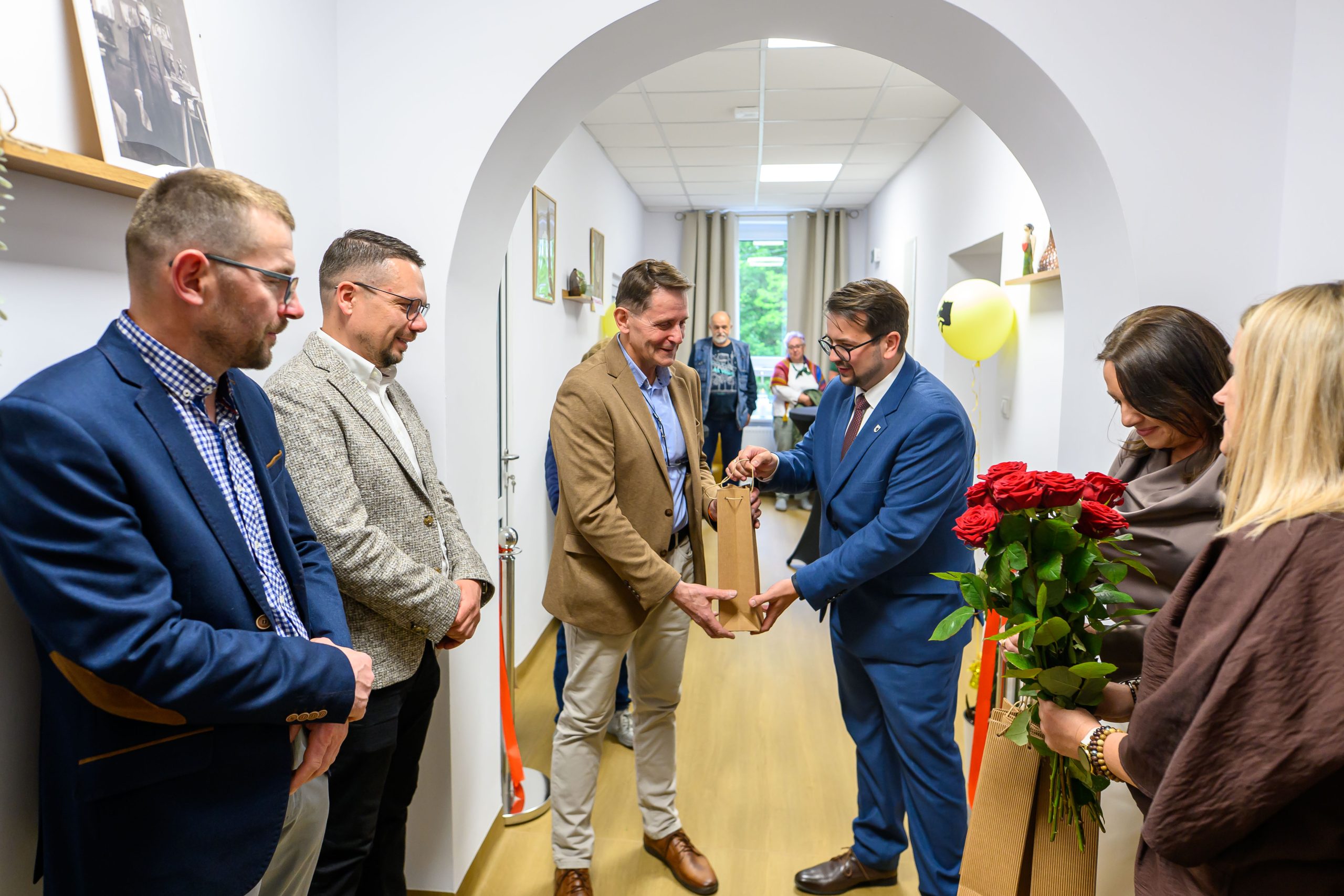 Grupa osób w garniturach i eleganckiej odzieży stoi w jasnym korytarzu. Jeden z mężczyzn przekazuje prezent drugiemu. Po prawej kobieta trzyma bukiet czerwonych róż.