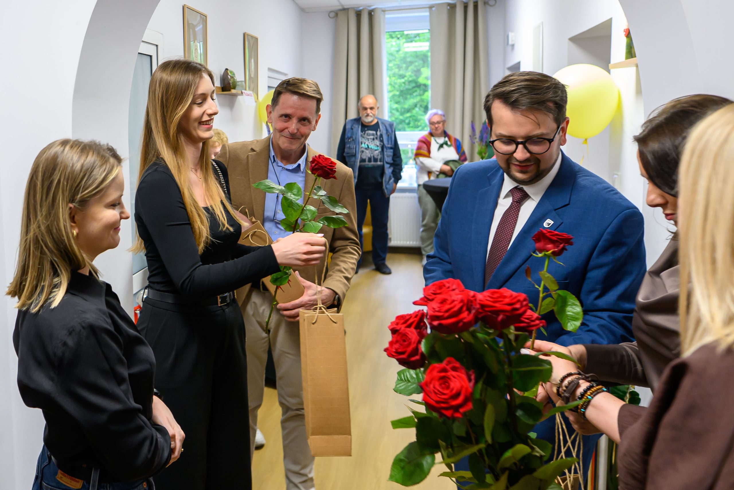 Grupa uśmiechniętych osób wymienia czerwone róże w jasnym pomieszczeniu. Tło z białymi ścianami i jasnymi zasłonami. Obecne elementy dekoracyjne i balon.