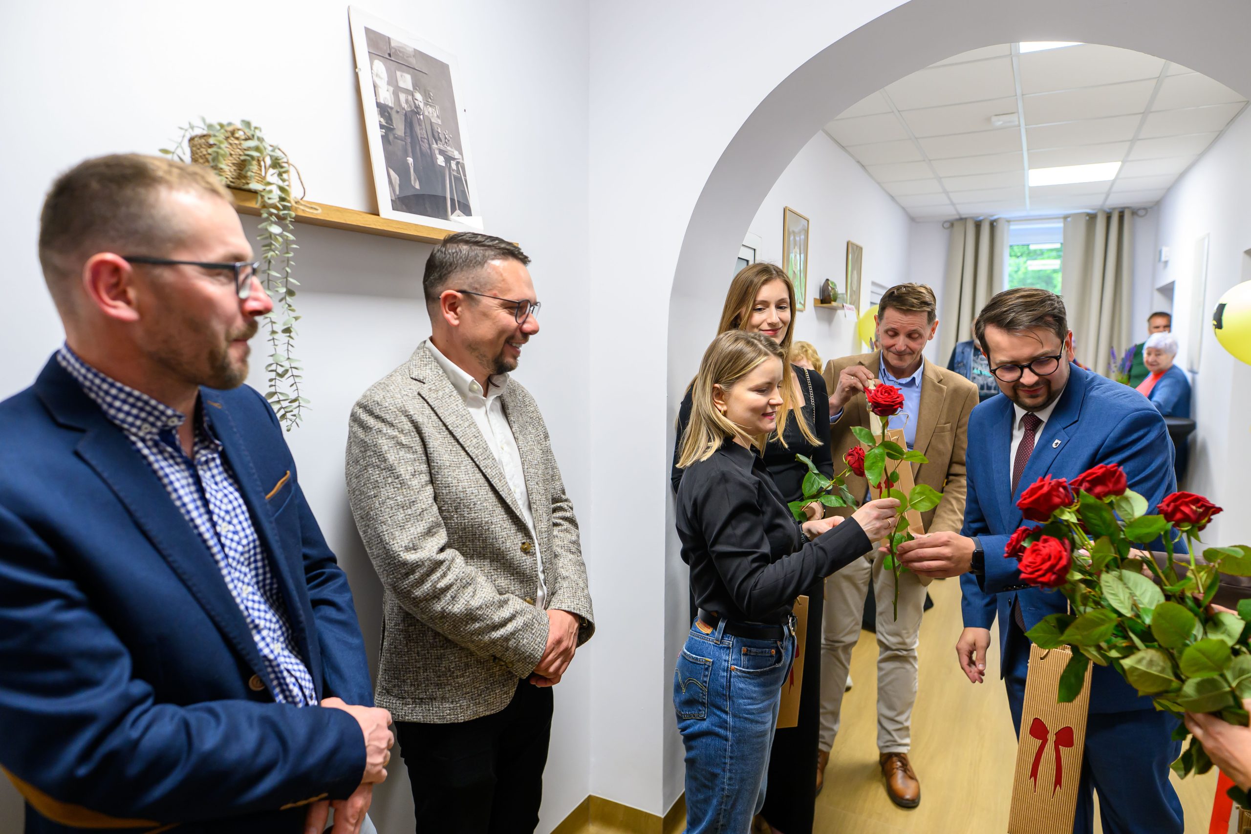 Grupa ludzi w eleganckich strojach rozdaje czerwone róże w jasnym pomieszczeniu. Na ścianie wiszą obrazy i półka z rośliną. Tło jest jasne z żółtymi balonami.