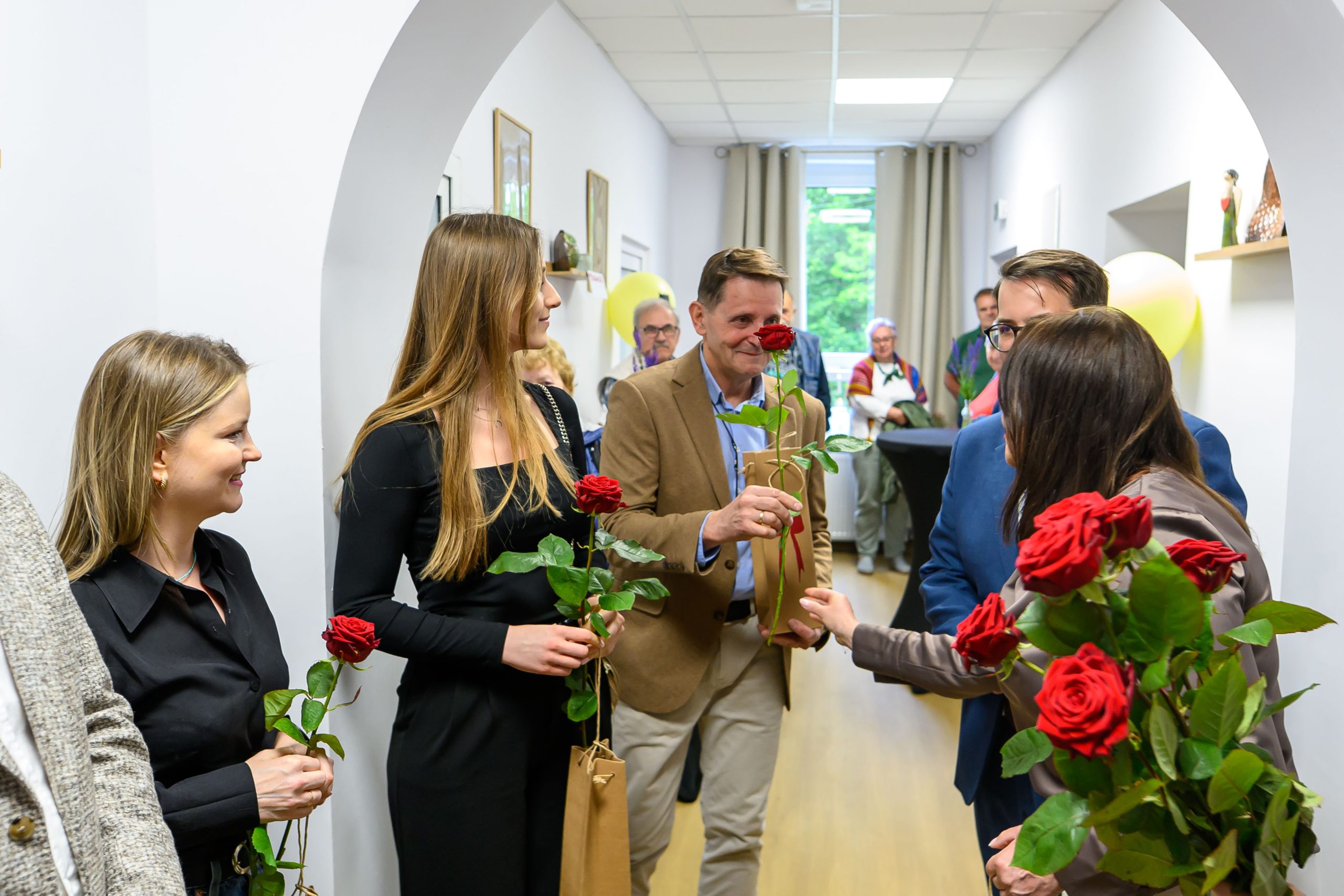 Grupa osób stoi w korytarzu z białymi ścianami, trzymając czerwone róże. Kobiety w czerni, mężczyzna w brązowej marynarce. W tle widać okno i balon.
