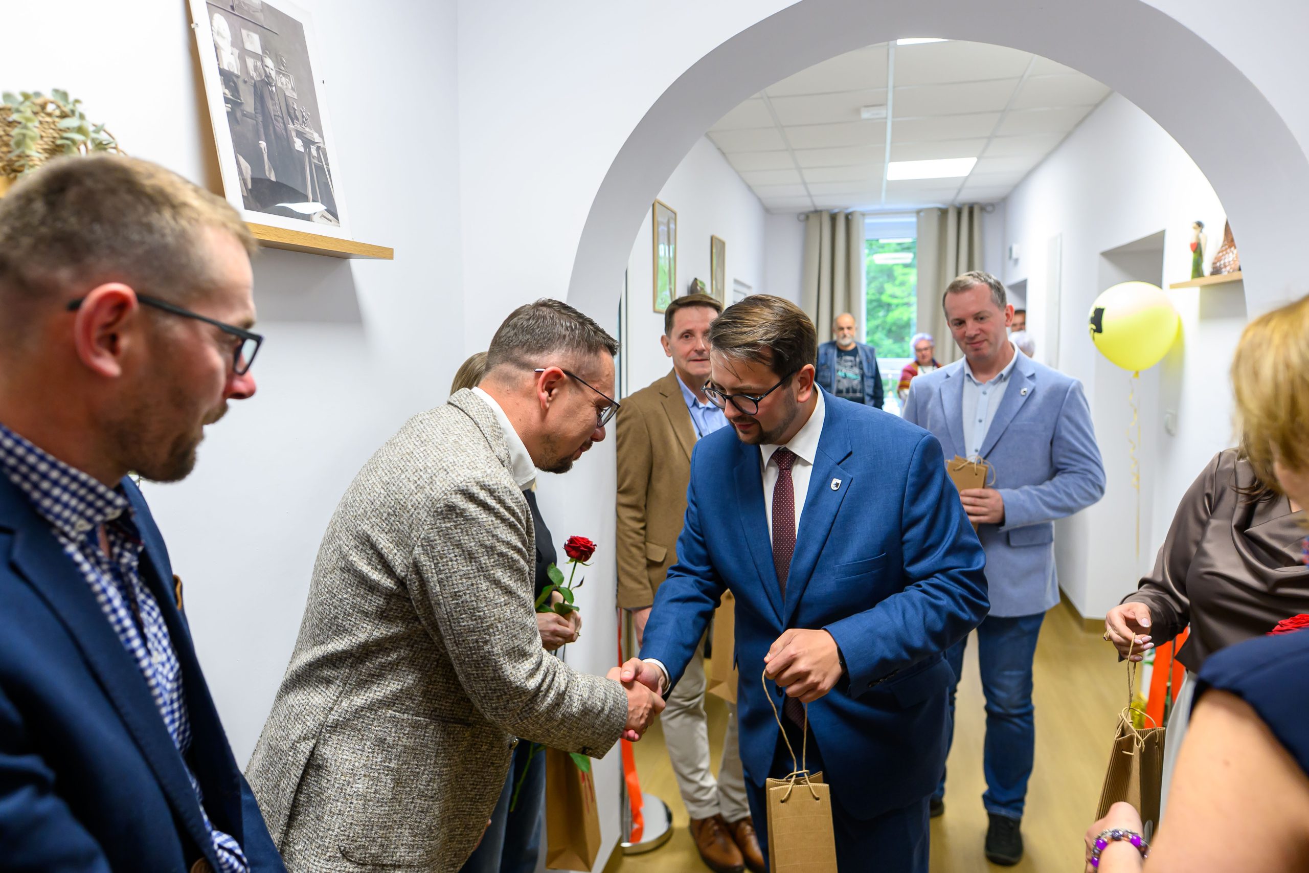 Grupa ludzi w eleganckich strojach w jasnym korytarzu, mężczyzna w granatowym garniturze wita się z osobą w szarej marynarce. Obrazy wiszą na białych ścianach. Balon i prezenty dodają uroczystego charakteru