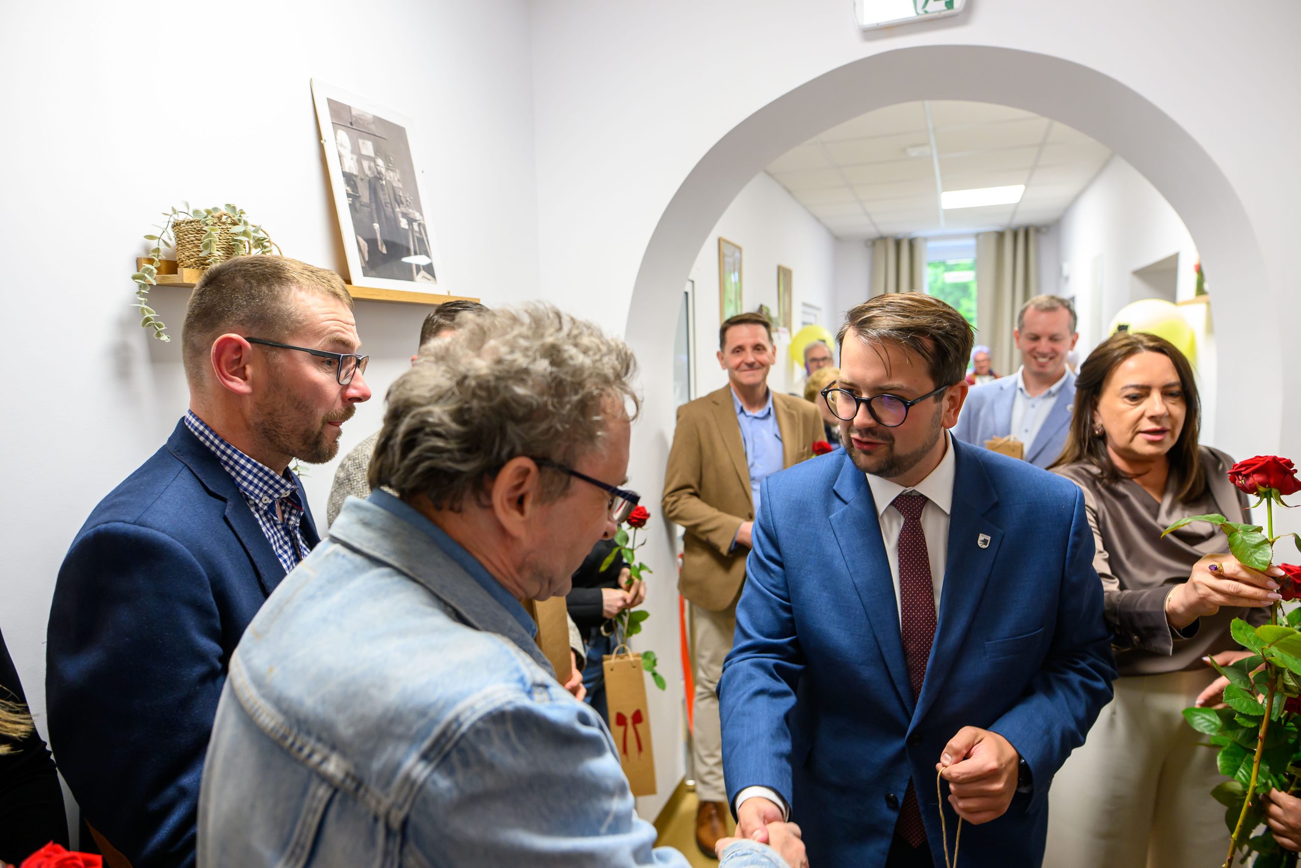 Grupa ludzi w eleganckiej odzieży w jasnym pomieszczeniu; jeden mężczyzna w niebieskim garniturze ściska rękę innemu. W tle białe ściany i obrazy. Kobieta trzyma czerwone róże.