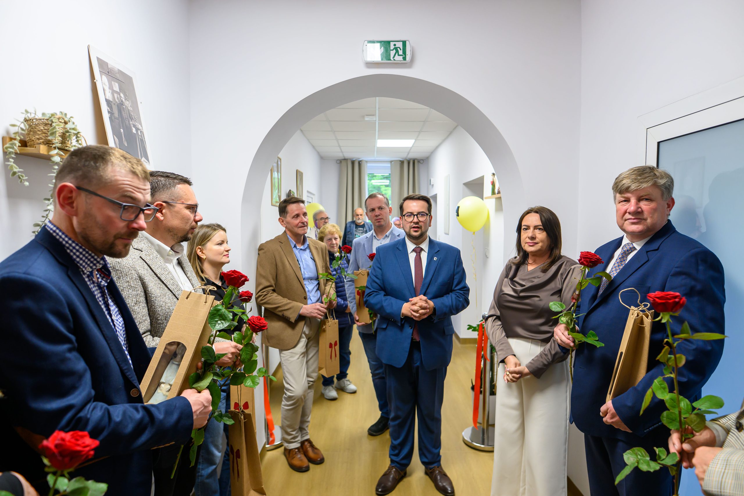 Grupa ludzi w formalnych ubraniach stoi w jasnym korytarzu. Większość trzyma czerwone róże i torebki. Ściany są białe, a podłoga jasnobrązowa.